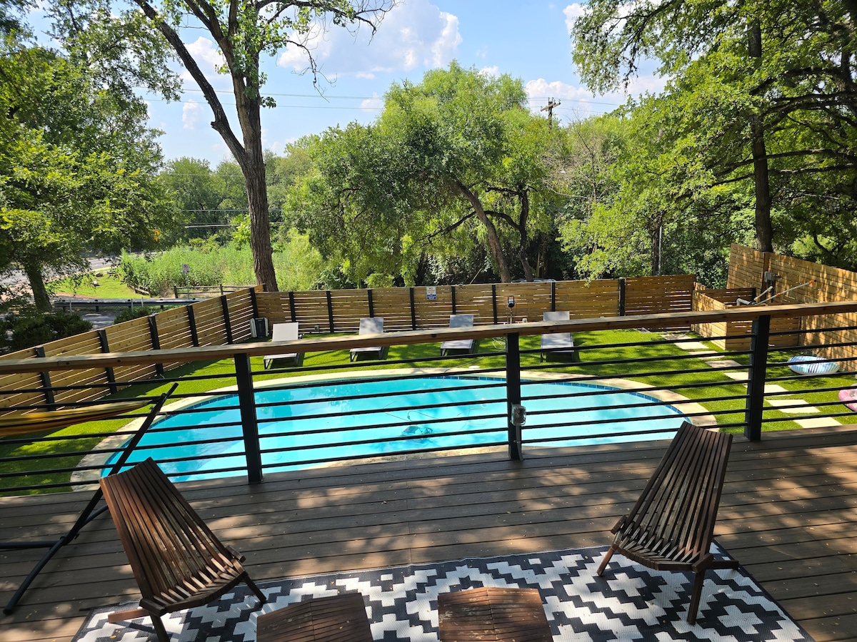 A private pool is surrounded by a fenced-in yard featuring lush green grass. Comfortable lounge chairs are positioned on a wooden deck, offering a view of the pool and the natural landscape beyond. The area is enhanced by shaded trees and a wooden railing.