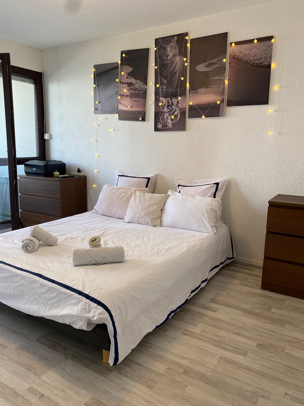 A bedroom is presented with a spacious bed dressed in white linens and accented with navy trim. Decorative cushions are arranged at the head of the bed. Above, framed artwork is displayed on the wall, enhanced by softly glowing string lights, adding a cozy touch.
