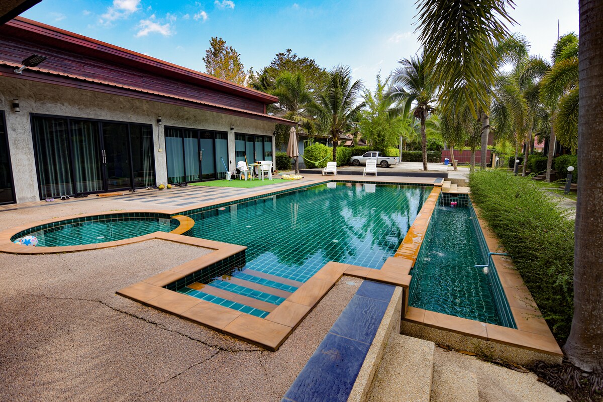 A shared swimming pool is surrounded by lush greenery, featuring turquoise tiles and a separate area for relaxation. Sun loungers and a parasol are positioned nearby, providing a serene space to unwind. Palm trees gently sway in the background, contributing to the peaceful environment.