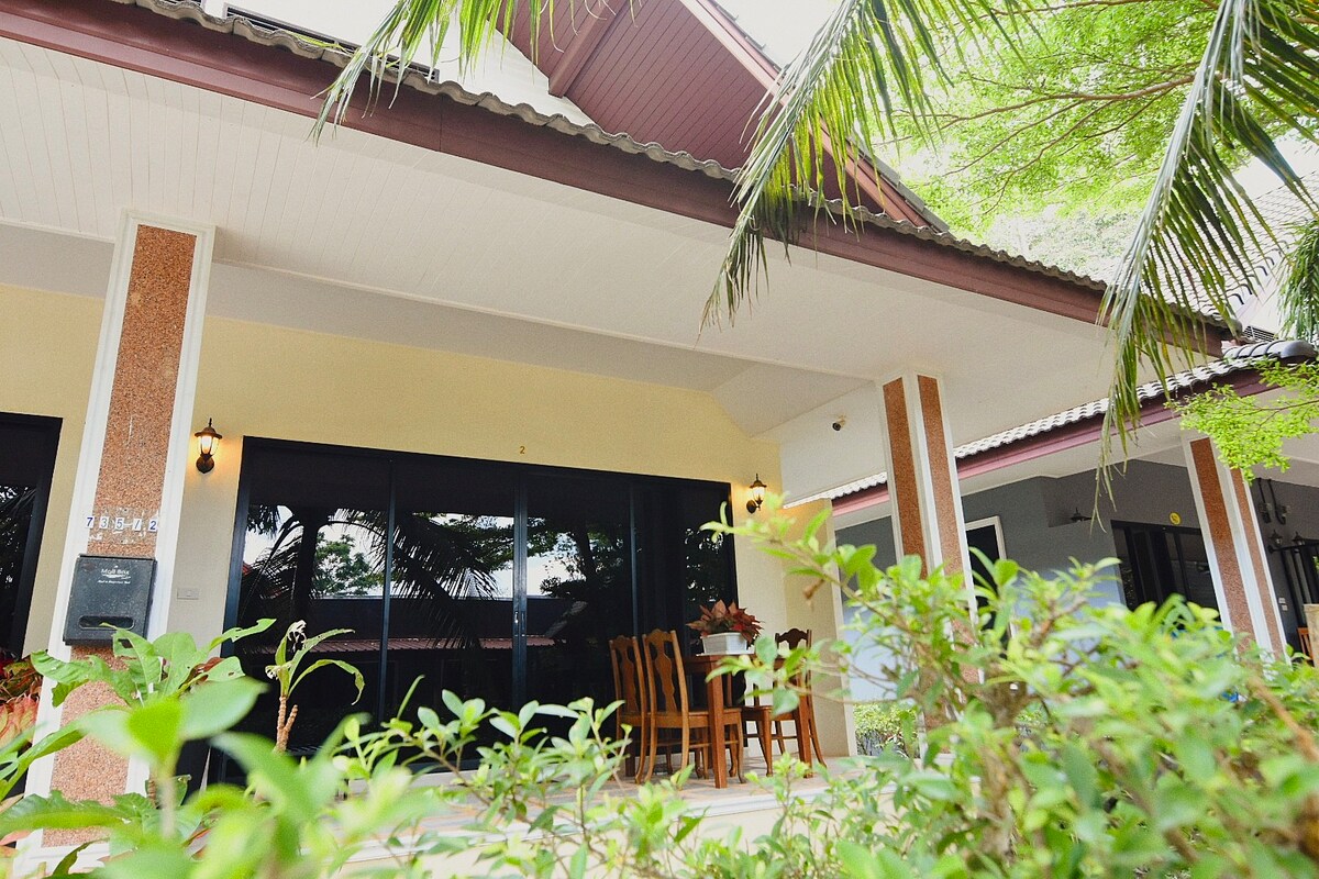 The villa's exterior is shown with a covered porch featuring a dining table and chairs. Lush greenery surrounds the entrance, complemented by a backdrop of palm trees. Large glass doors provide a view into the indoor space, inviting natural light into the home.