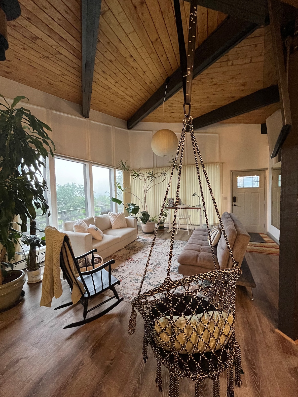 An airy living room features a high wood-paneled ceiling and large windows that offer views of the surrounding greenery. A swinging chair adds a unique touch, while a comfortable sofa and potted plants enhance the tranquil ambiance of the space.