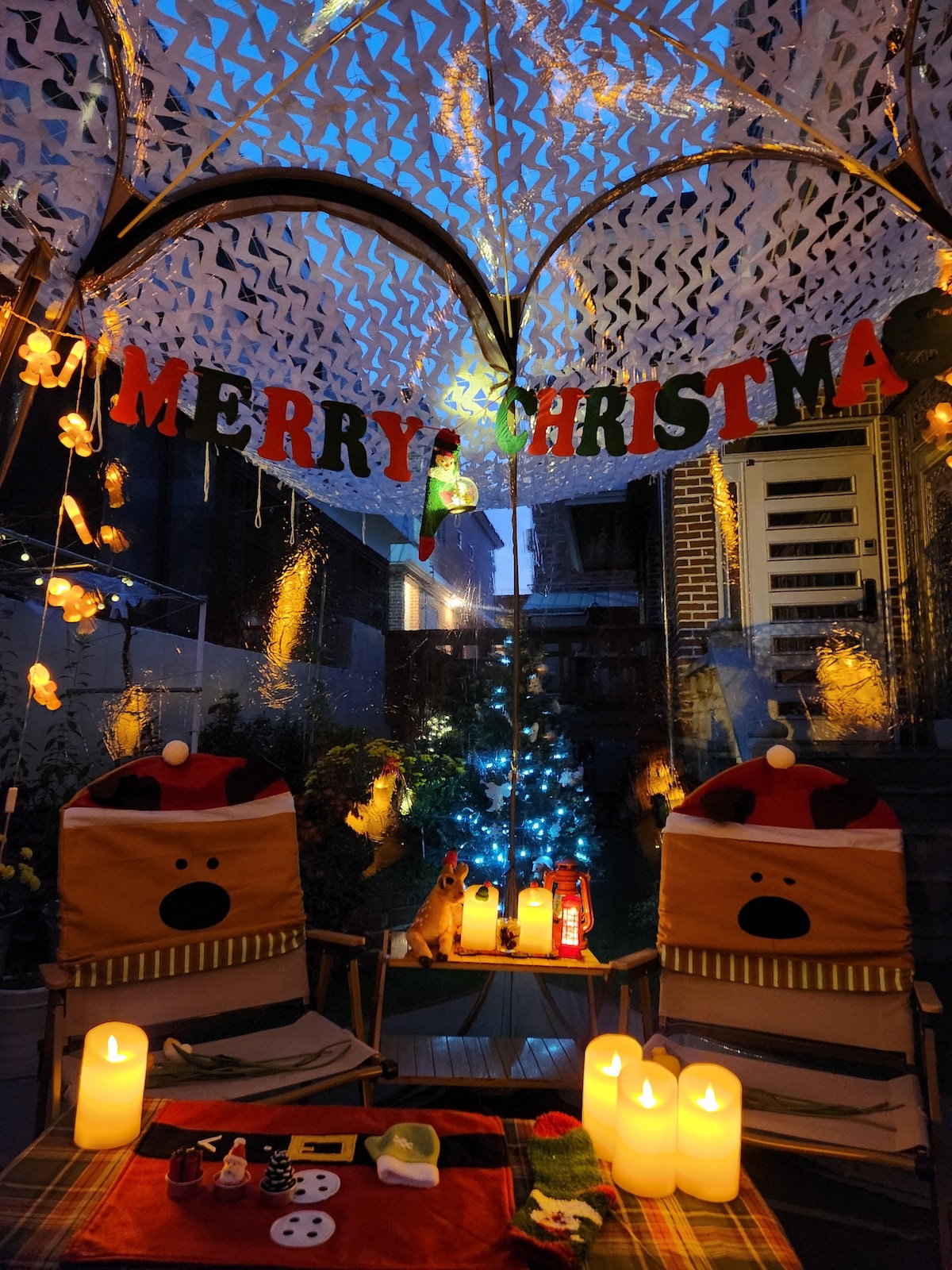 An outdoor area is decorated with holiday-themed decor, including a 'MERRY CHRISTMAS' banner and a small tree with lights. Two festive chairs embellished with animal designs are positioned around a table set with candles and various festive items, creating a cozy atmosphere.