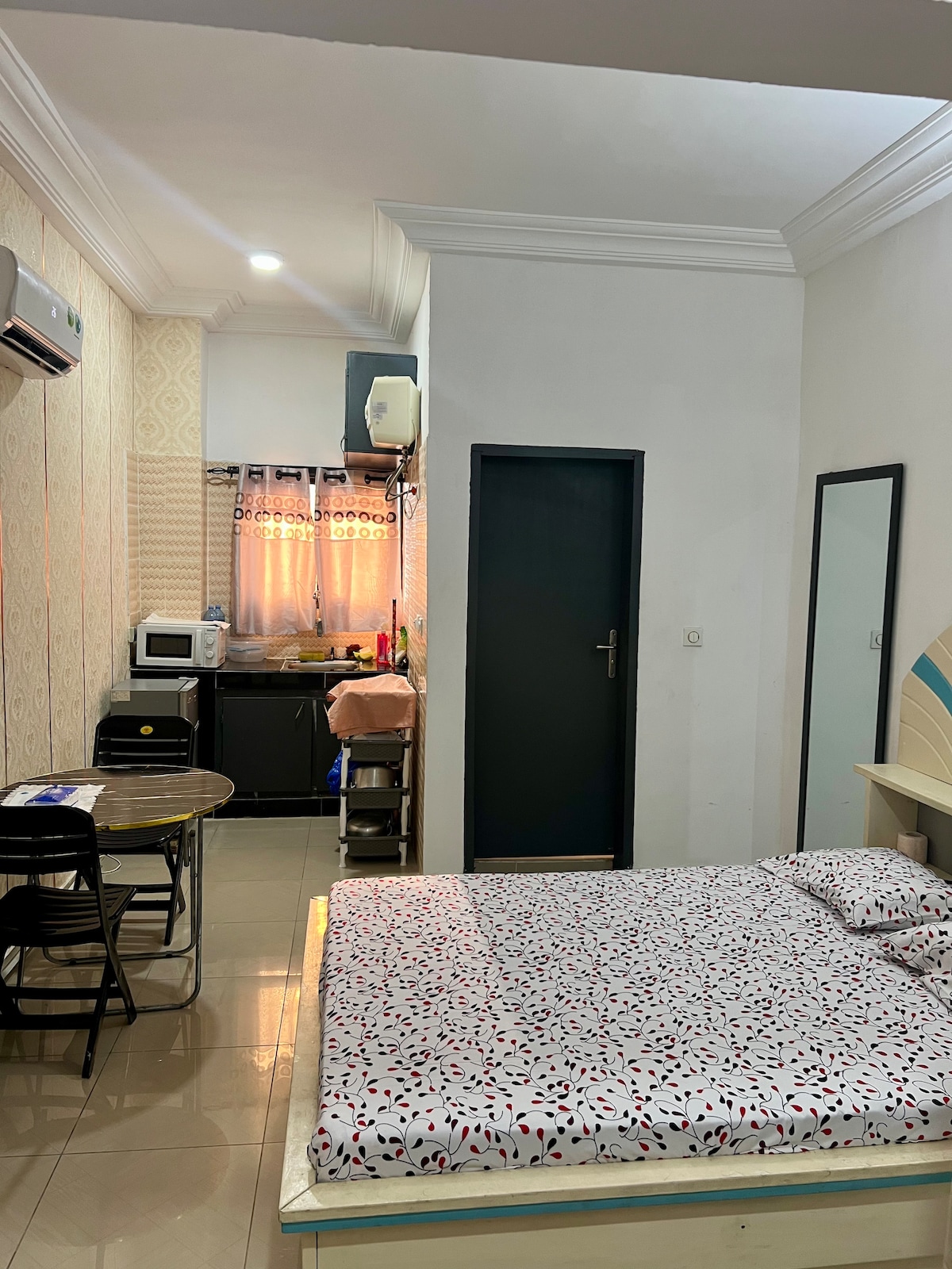A well-arranged studio features a spacious sleeping area with patterned bedding. A kitchenette is visible in the corner, equipped with necessary appliances. A dining area with chairs is included, and ample natural light enters through the window adorned with light curtains.