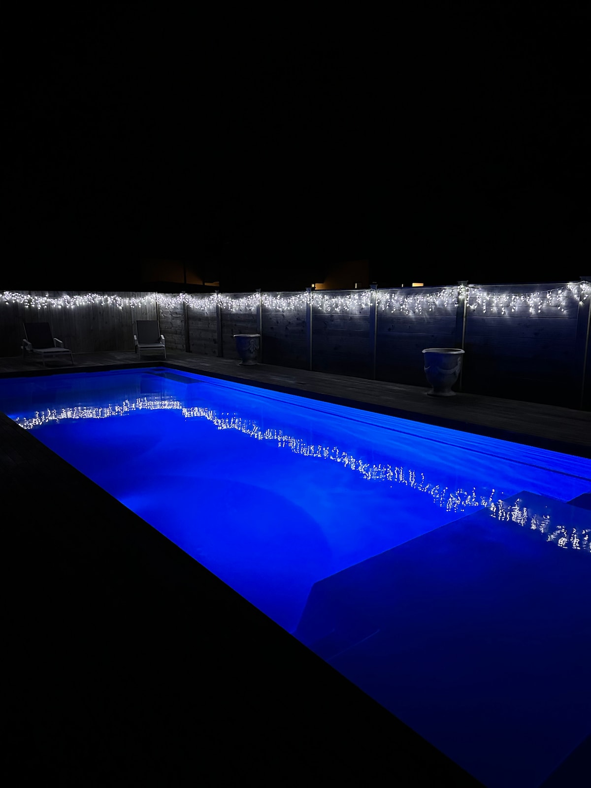 A saltwater pool is illuminated in a calming blue hue, reflecting the surrounding lights. Decorative string lights are seen lining the enclosure, enhancing the tranquil ambiance during the night.