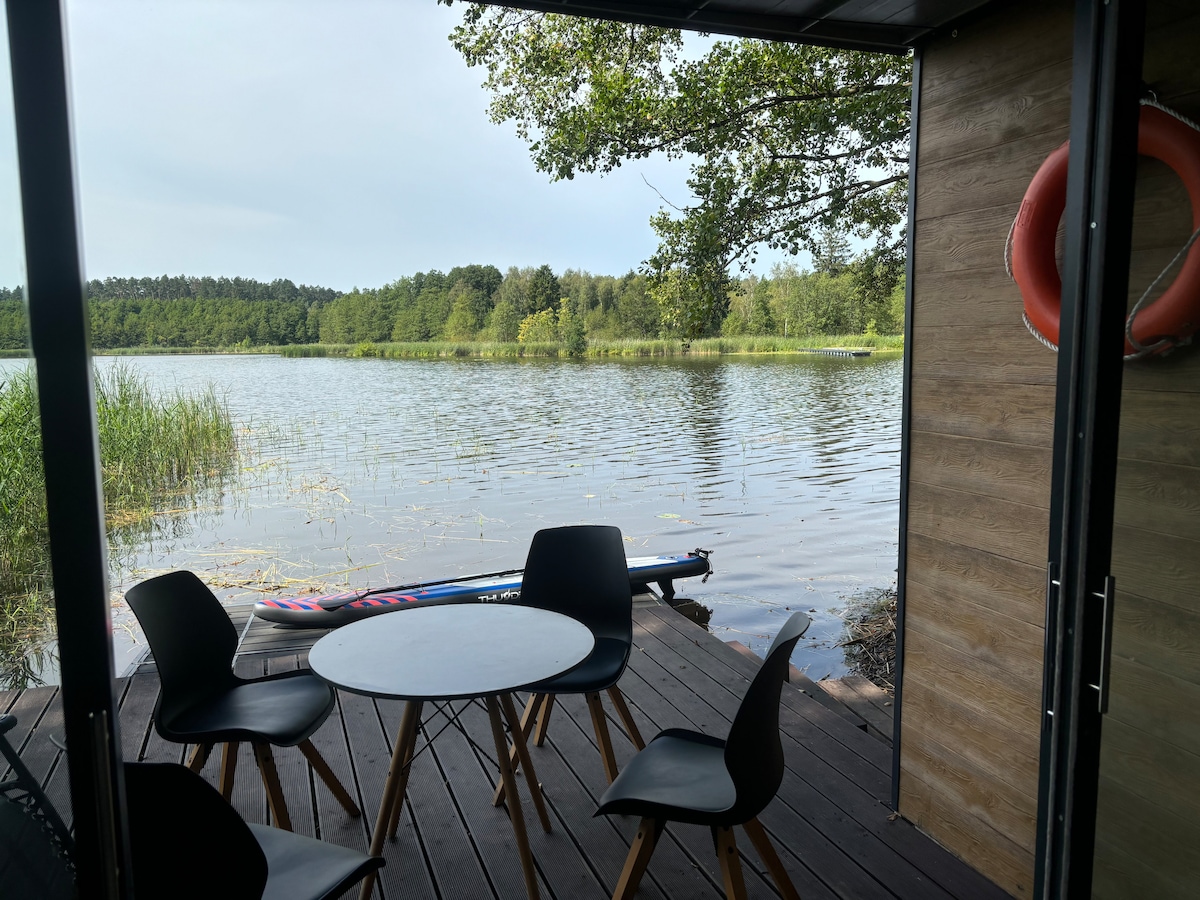 A wooden deck extends from the cabin towards the lake, featuring a small round table surrounded by four chairs. In the background, the calm water reflects the greenery along the shoreline, while a paddleboard rests on the edge of the lake.