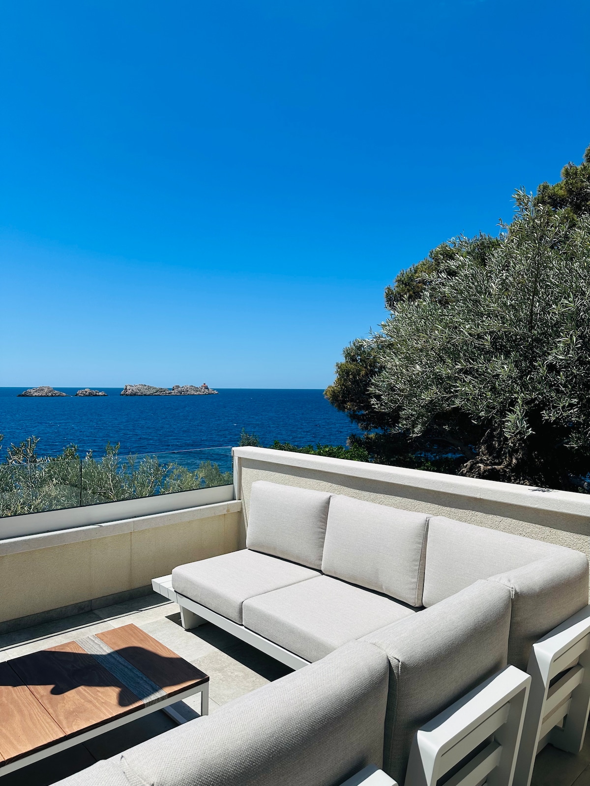 A spacious outdoor seating area is presented, featuring a light-colored sofa set positioned near the edge of the terrace. The view overlooks the calm sea and distant rocky formations under a clear blue sky, surrounded by greenery.