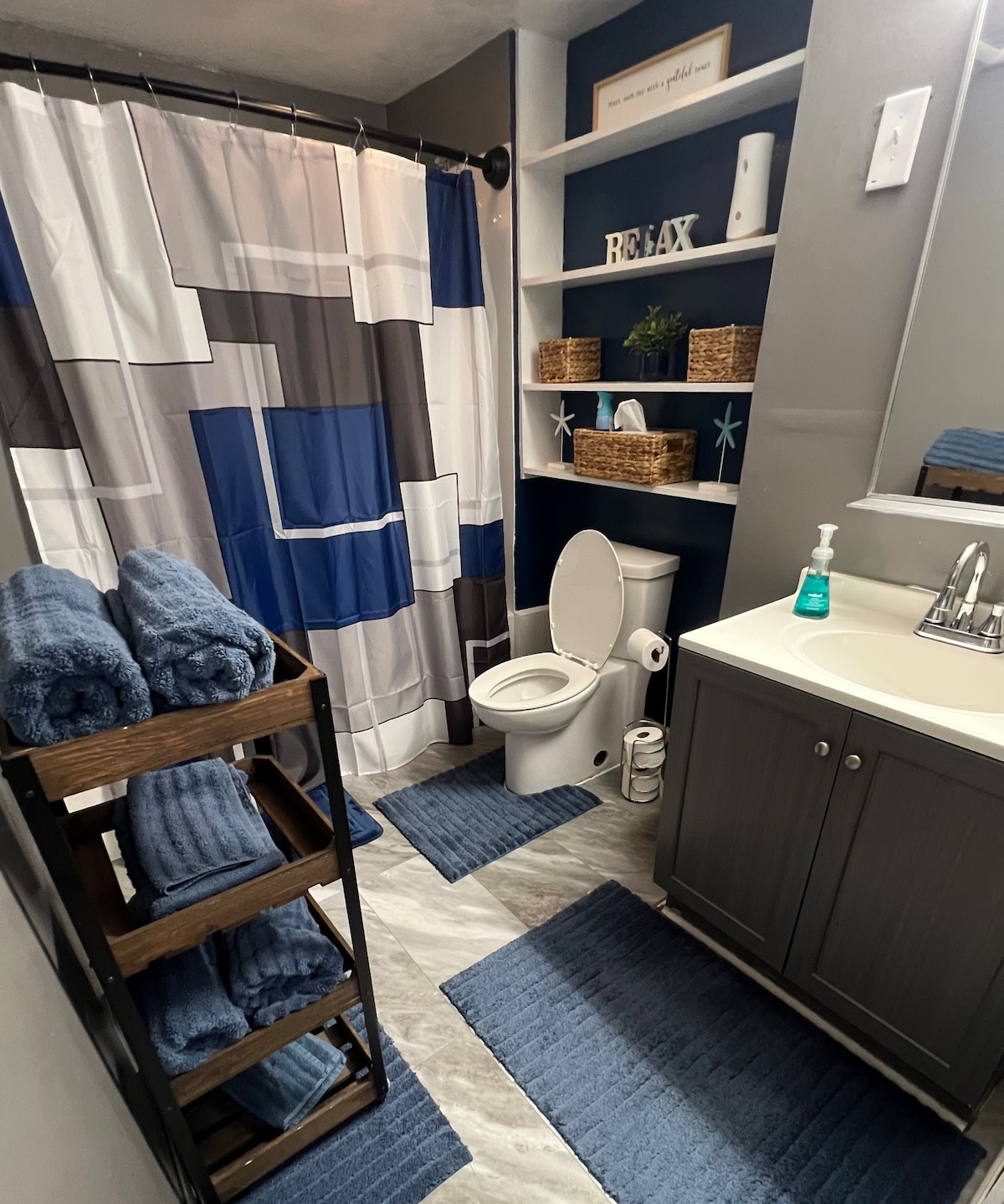 A neatly arranged bathroom features a blue and grey shower curtain alongside a white toilet. A wooden rack displays rolled towels, and a small shelf holds decorative items. The flooring exhibits a light marble pattern, while plush bath mats complement the overall color scheme.