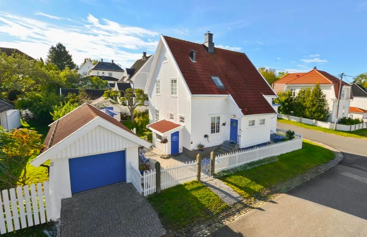 An elevated view of a charming white villa with a red tiled roof, set in a residential area. The property features a well-maintained yard with greenery and a white picket fence. A driveway leads to a blue garage, contributing to the welcoming exterior.