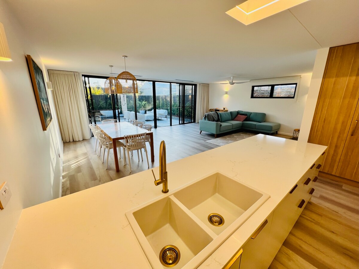 An open-plan living area features a modern kitchen with dual sinks, leading to a spacious dining table and comfortable seating area. Large glass doors open to an outdoor terrace, allowing natural light to fill the room. Light wood flooring complements the neutral color palette.