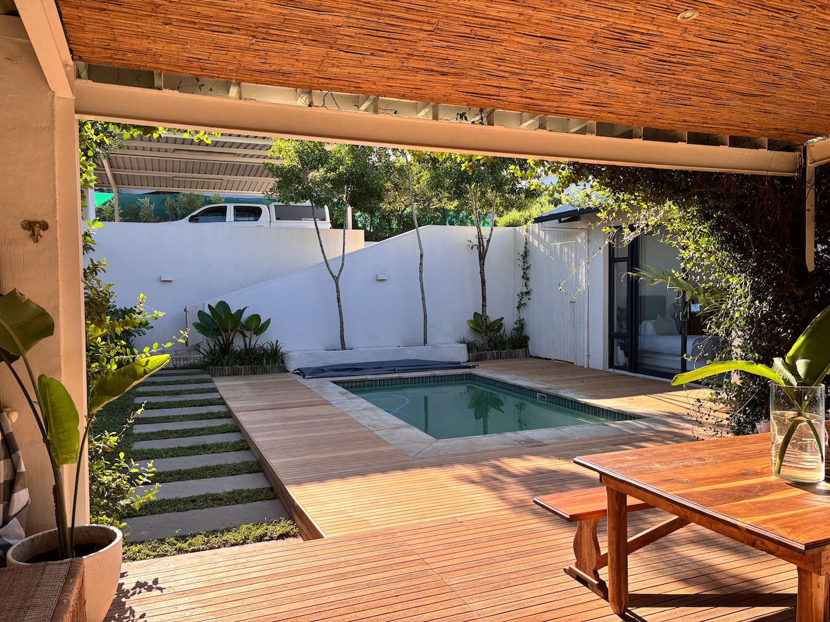 The image shows a private outdoor area with a pool surrounded by wooden decking. Lush green plants are present, enhancing the tranquil feel. The space is shaded under a bamboo roof, providing a comfortable environment. A wooden table is placed in the foreground, ideal for relaxation.