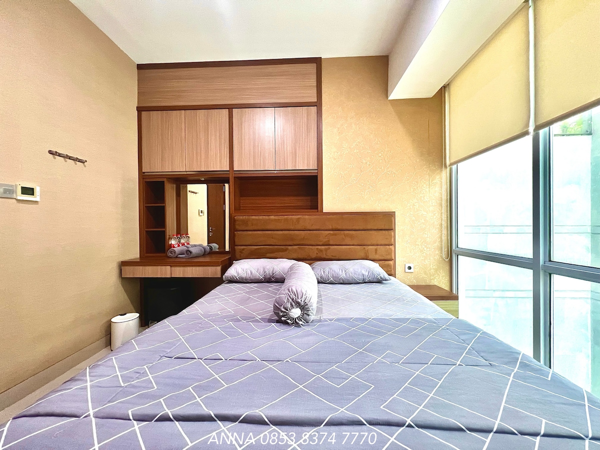 A cozy sleeping area features a spacious bed with a light grey patterned blanket. Nearby, a wooden headboard complements built-in shelving and a vanity mirror. Natural light enters through large windows with sleek shades, creating a peaceful ambiance.