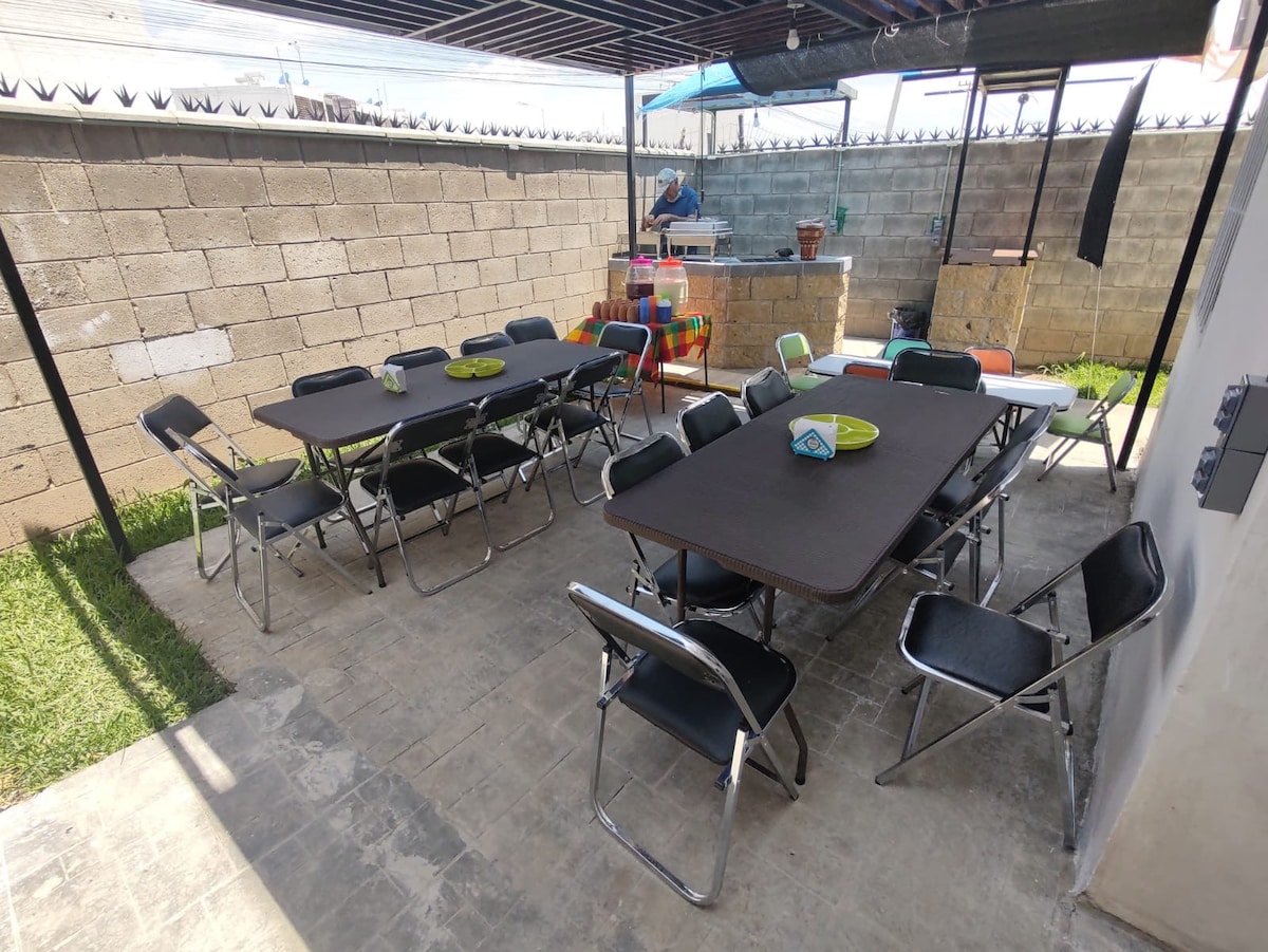 A spacious outdoor area features multiple folding tables and chairs arranged for gatherings. The ground is paved, and vibrant green grass is visible along the edges. A grill and colorful containers are seen in the background, surrounded by a block wall providing privacy.
