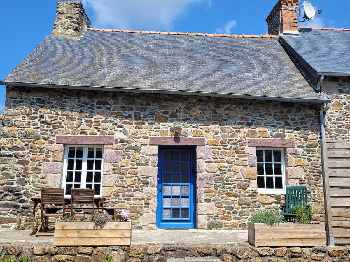 A rustic stone cottage is displayed with a blue door as the central feature. Two windows flank the entrance, while a small patio area includes wooden chairs and planters. The structure is set against a clear blue sky, enhancing the tranquil ambiance.