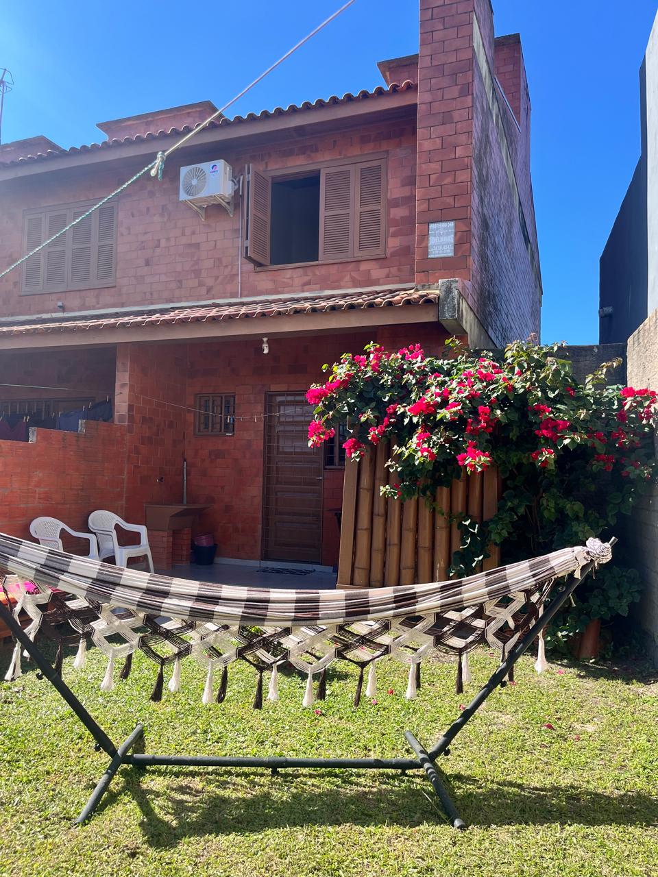 A two-story brick home is visible, featuring wooden window shutters and an air conditioning unit. In the foreground, a hammock is gently draped over a stand. Colorful flowers cascade from a nearby wall, enhancing the inviting outdoor space surrounded by grass.
