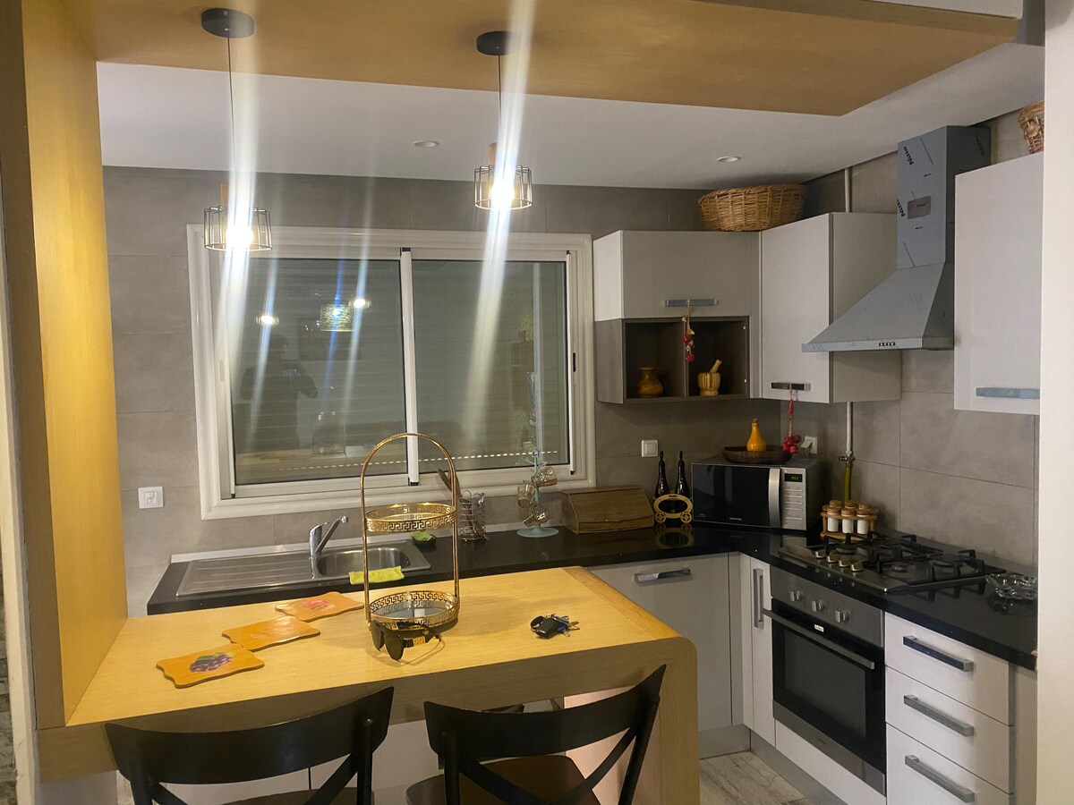 The modern kitchen features a functional layout with a wooden island, black bar stools, and stainless steel appliances. Cabinets in neutral tones provide ample storage. Under-cabinet lighting illuminates the workspace, while a large window allows natural light to brighten the area.