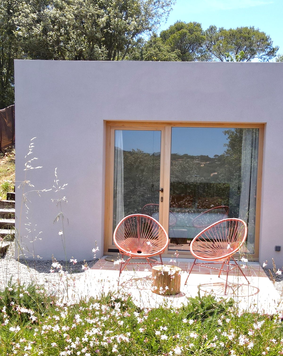 A private patio features two coral-colored chairs facing large glass doors. The surrounding area is composed of soft grasses and wildflowers, providing a natural setting. Behind the doors, a calm interior space is hinted at, drawing attention to the connection between indoor and outdoor areas.