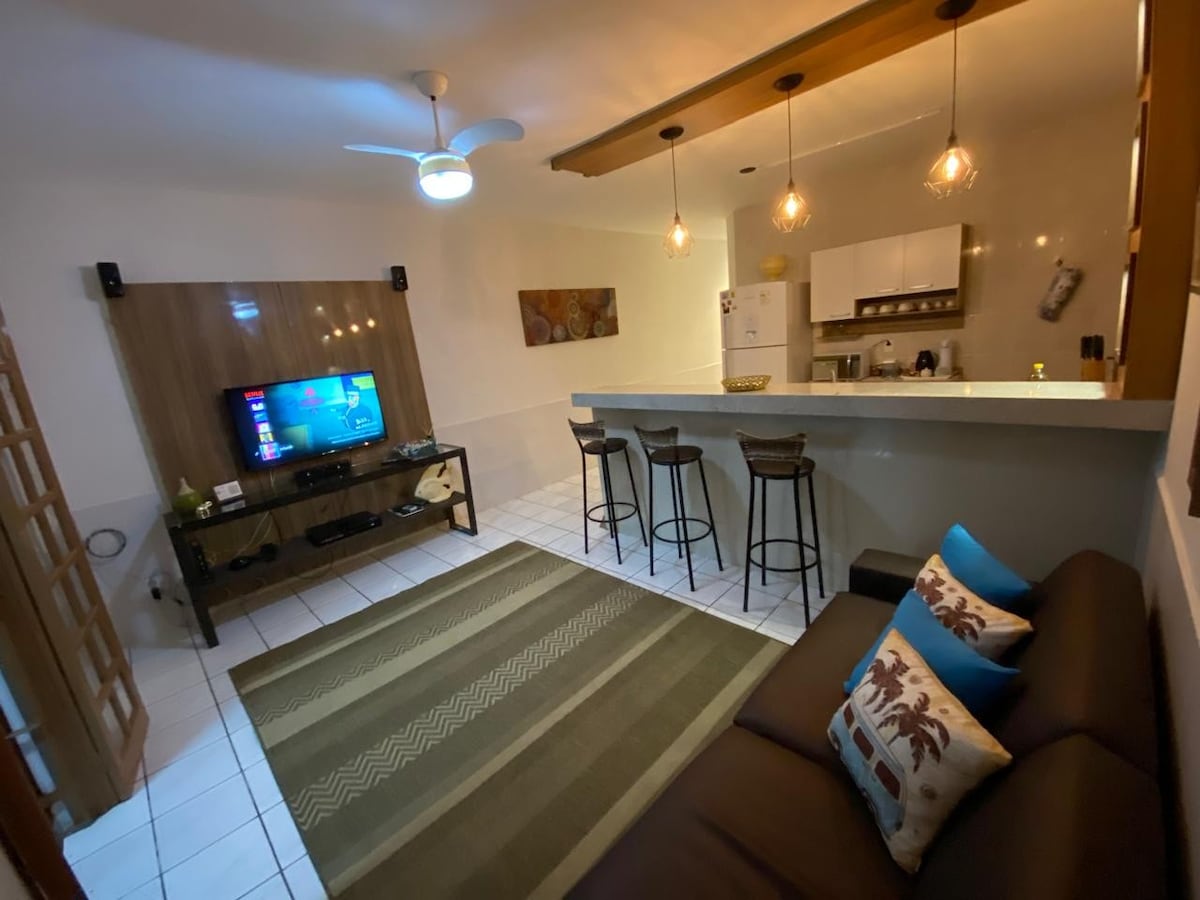 The living area features a comfortable sofa adorned with decorative pillows, facing a television on a low table. A modern kitchen bar with three stools is seen in the background. Soft lighting adds warmth, and a textured rug lies on the tiled floor.