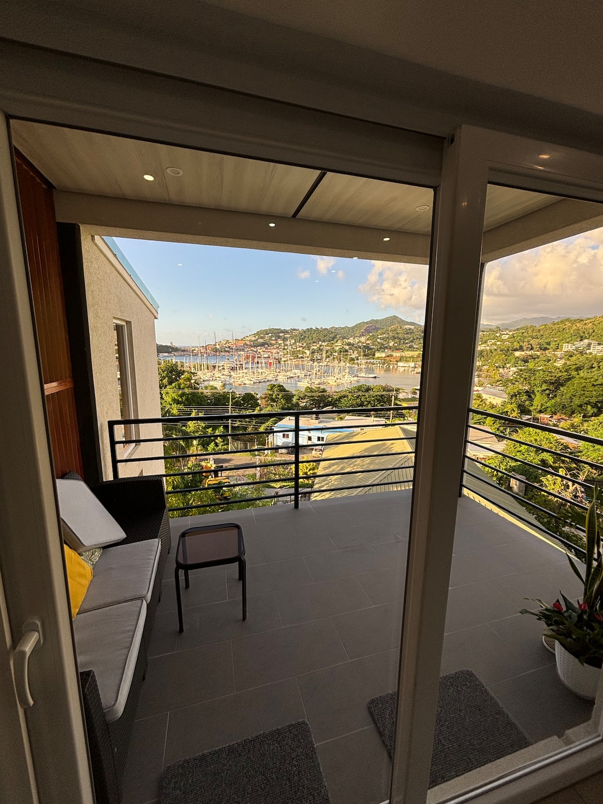 A balcony is shown with a seating area featuring a sofa and a chair. Glass doors open to a view of the lush landscape and water in the distance, overlooking the marina and surrounding hills. Natural light illuminates the outdoor space.