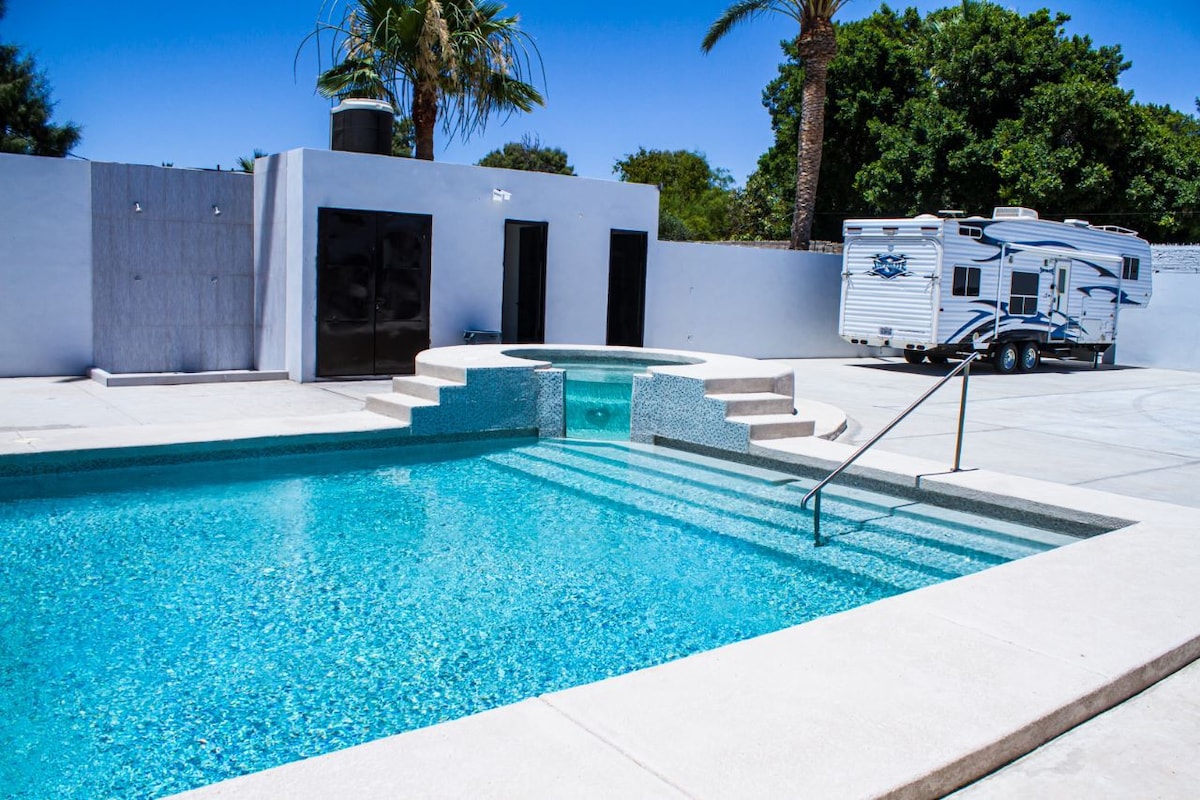 A bright and inviting swimming pool features clear turquoise water, with steps leading into it. A jacuzzi is positioned adjacent to the pool, surrounded by a spacious patio area. Palm trees provide shade in the background, with a trailer parked nearby.