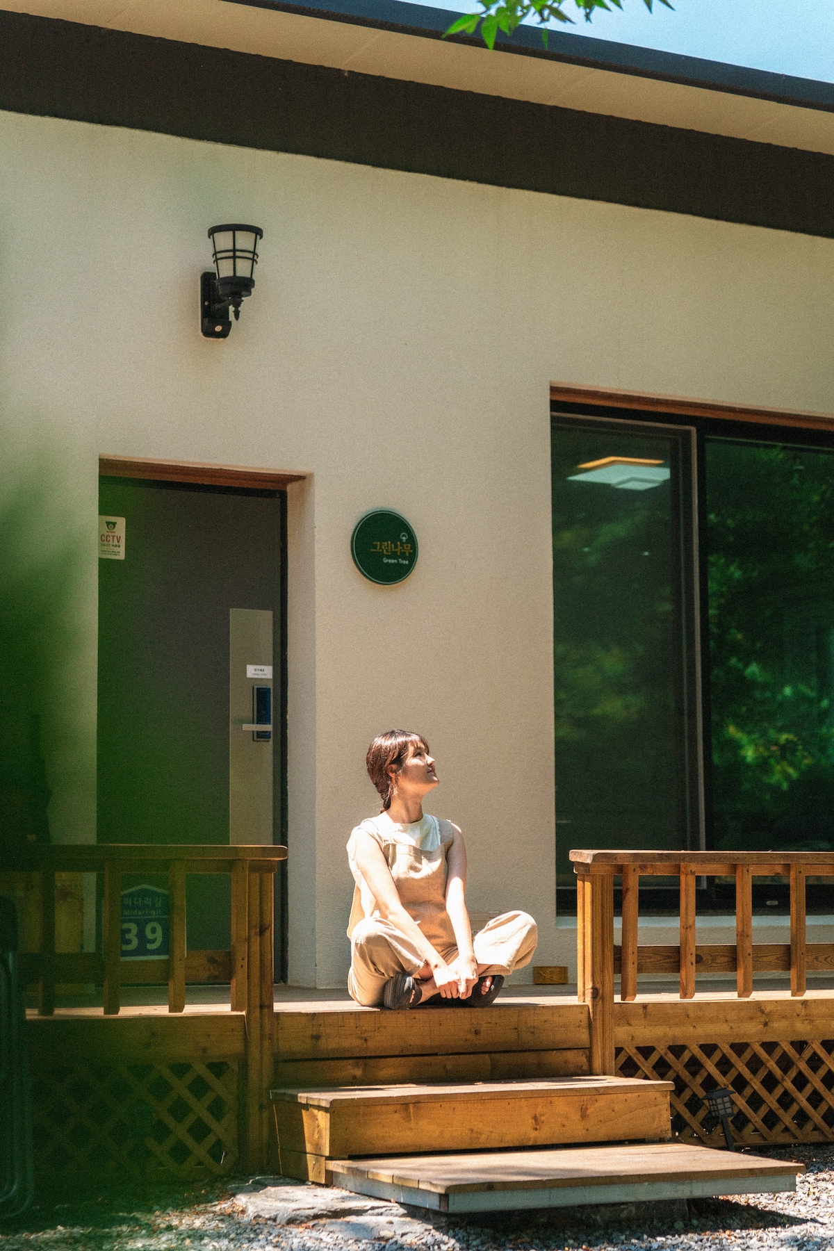 The entrance area is shown, featuring a wooden deck with steps. A person is seated, facing the door, surrounded by greenery. Large windows on the building allow for natural light, and a sign is visible near the door, indicating the unit number.