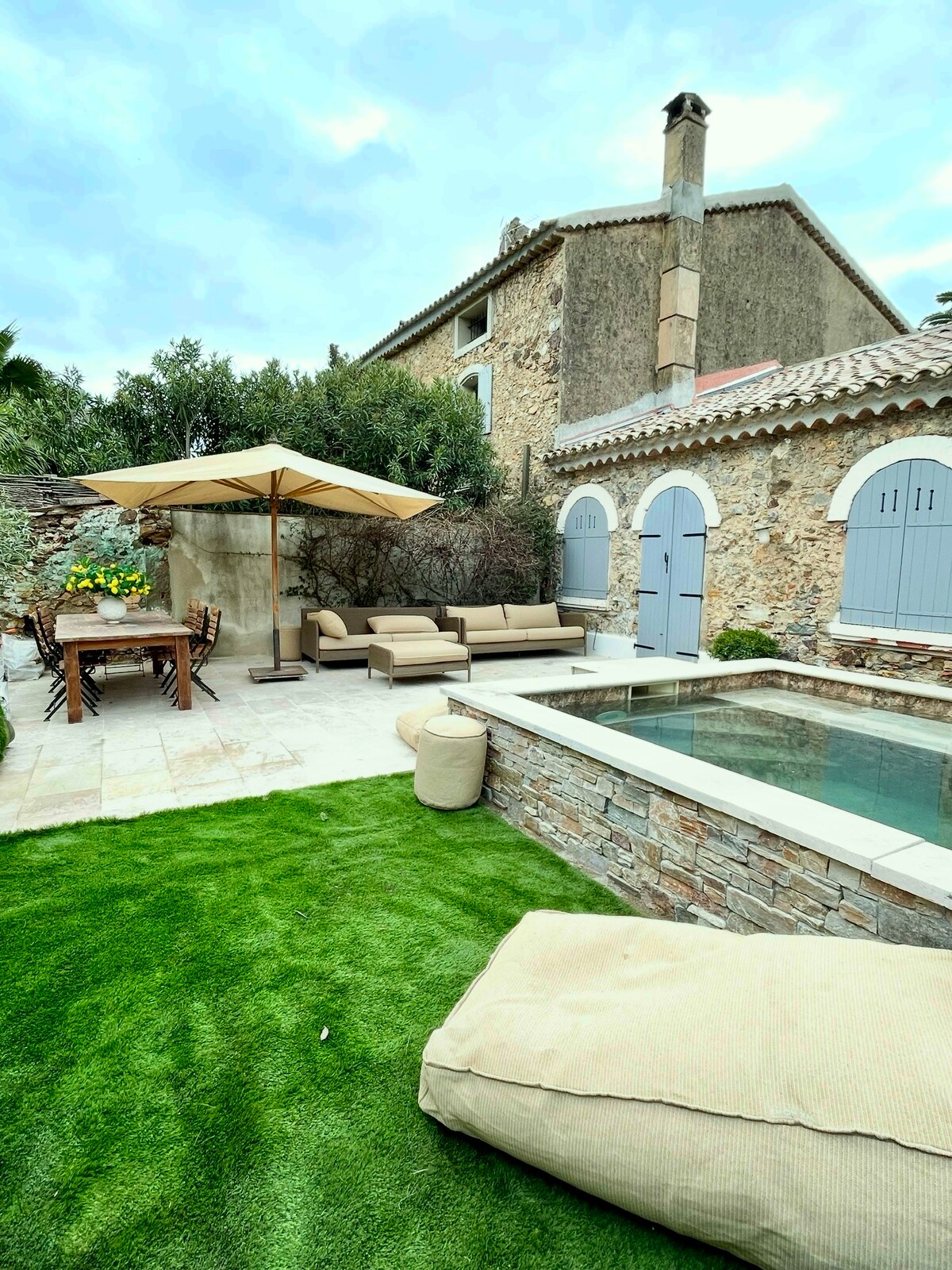 The outdoor area features a stone pool surrounded by green grass and lounge seating, under the shade of an umbrella. A wooden dining table is set nearby, offering a comfortable space for outdoor meals. The rustic architecture of the villa adds character to the space.