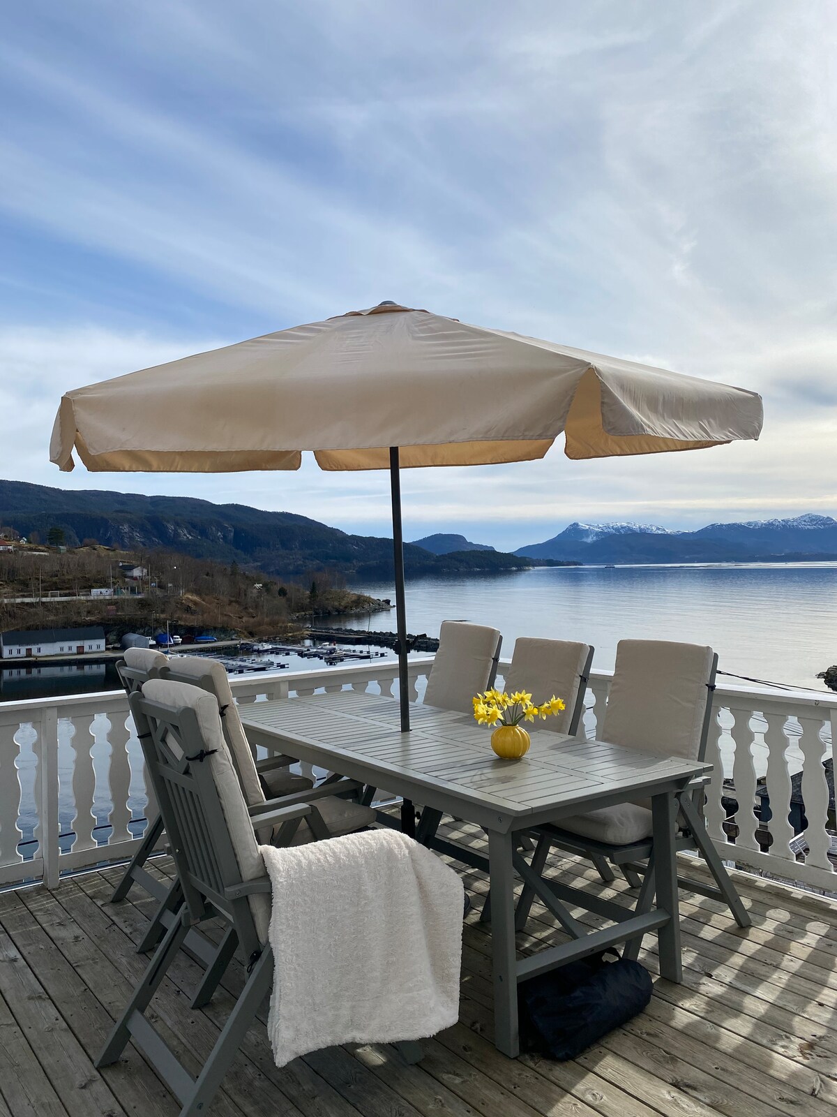 A spacious terrace features a wooden dining table surrounded by comfortable chairs, sheltered by a large umbrella. A vase with yellow flowers adds a touch of color, while the serene view of the fjord and mountains in the background creates a peaceful setting.