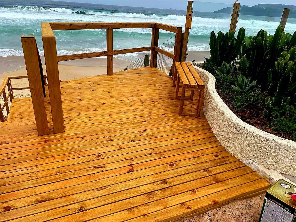 A wooden deck provides access to a sandy beach, surrounded by coastal vegetation. A small bench is visible on the deck, which offers a view of the ocean waves in the distance. Cacti are planted in a curved garden bed nearby.