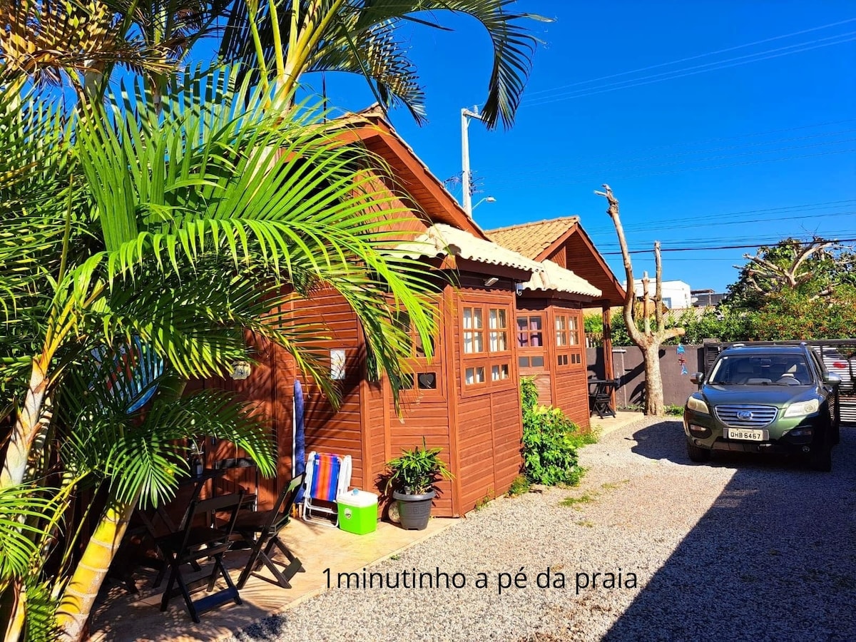 The exterior of the comfortable bungalows is shown, surrounded by lush green palm trees. A gravel pathway leads to the entrance, with a parked car visible nearby. The bungalows feature wooden siding and multiple windows, allowing for ample natural light.