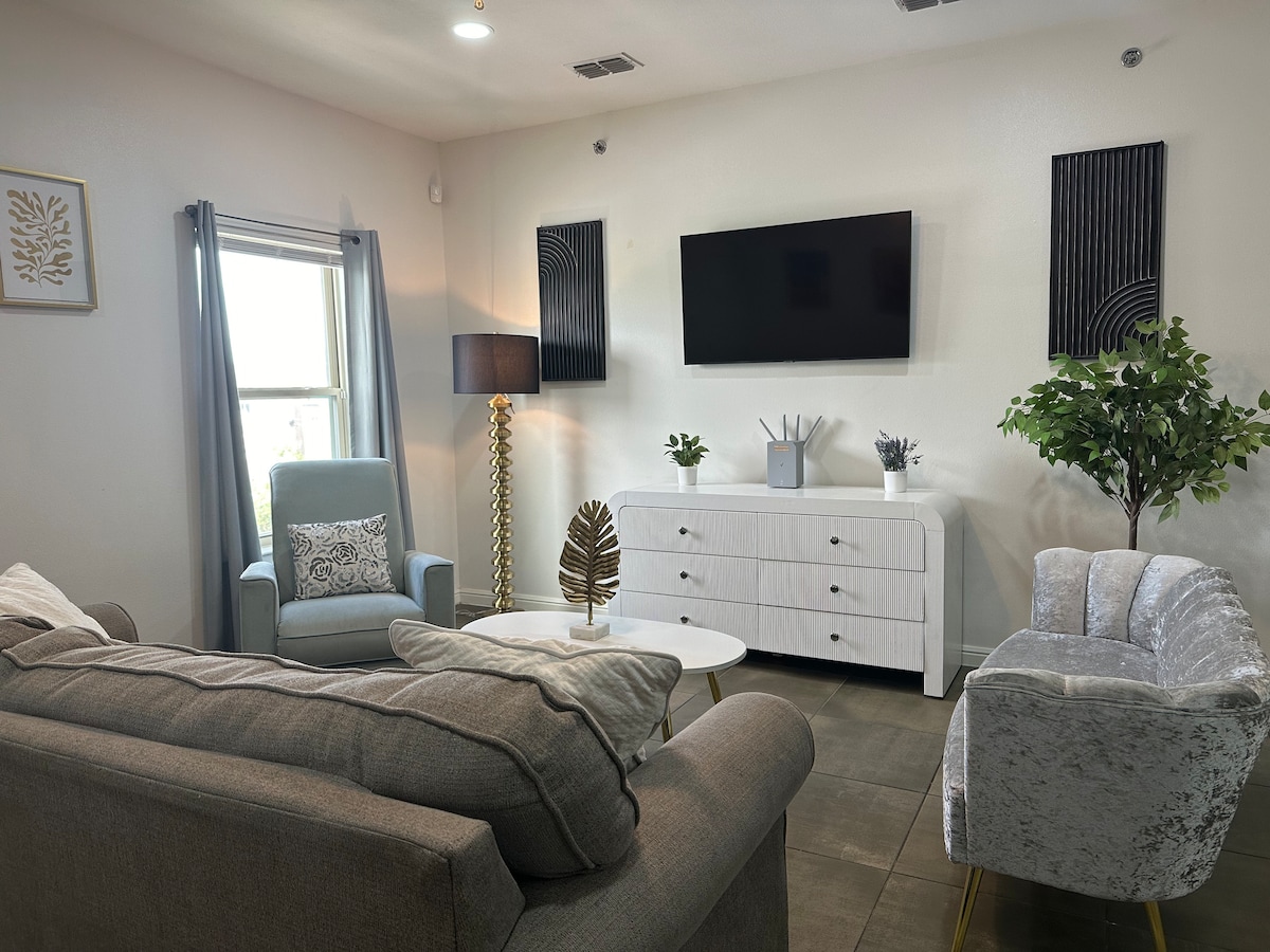 A welcoming living area is presented, featuring a gray sofa, two accent chairs, and a white coffee table. Modern decor is showcased with a large wall-mounted television and decorative plants. Natural light enters through a window with light curtains, enhancing the bright atmosphere.