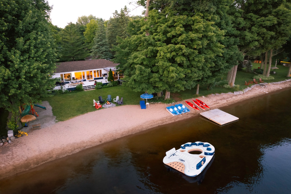 A serene beachfront scene showcases a sandy shore lined with trees. Colorful kayaks and paddleboats are arranged on the beach, while a floating dock extends into the tranquil water. A cozy cottage is visible in the background, framed by a spacious lawn and outdoor seating.