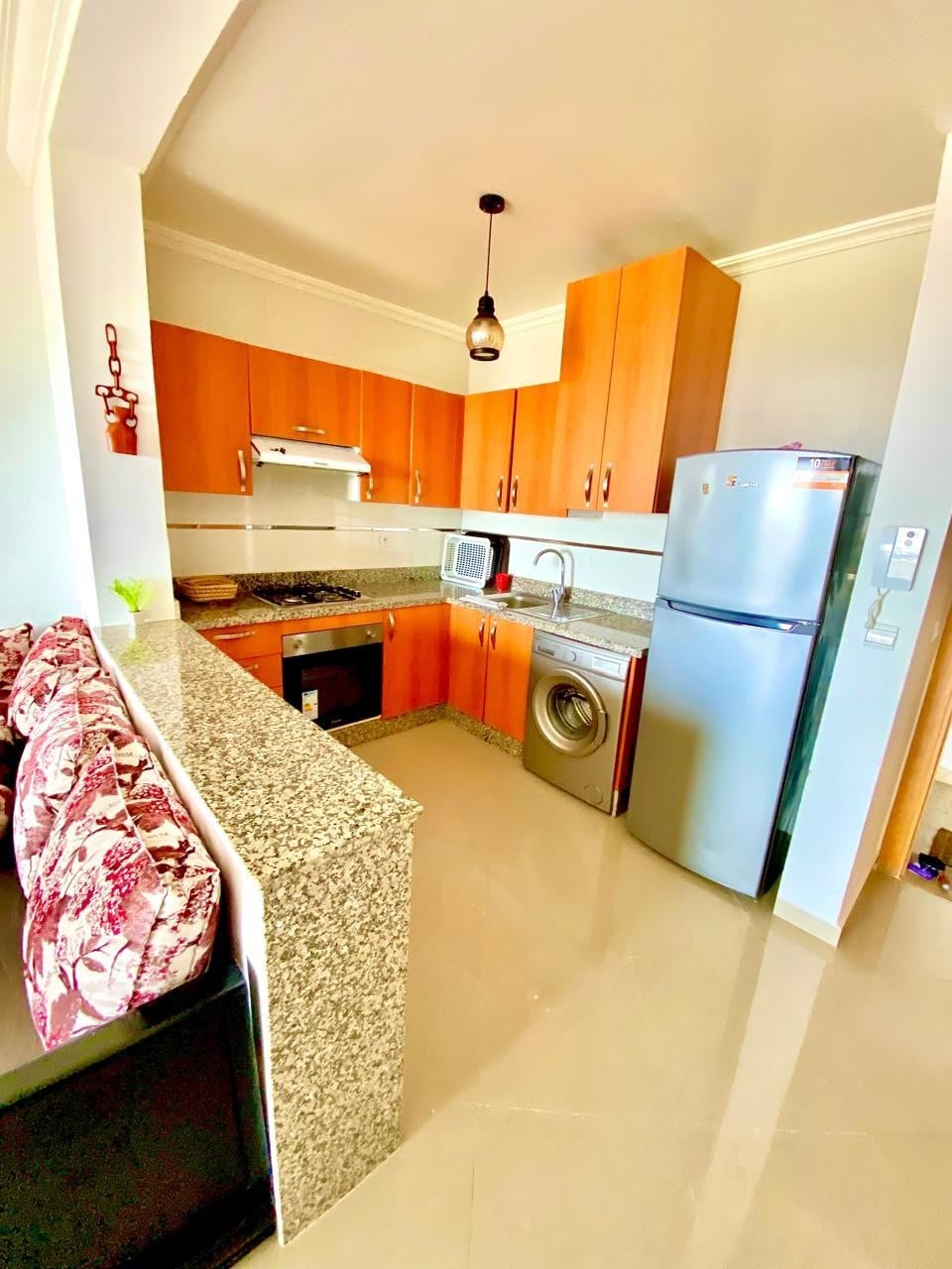 A modern kitchen is equipped with wooden cabinetry and stainless steel appliances, including a refrigerator and washing machine. A granite countertop offers ample workspace, while a small seating area is featured along one side with patterned cushions, adding comfort to the functional space.