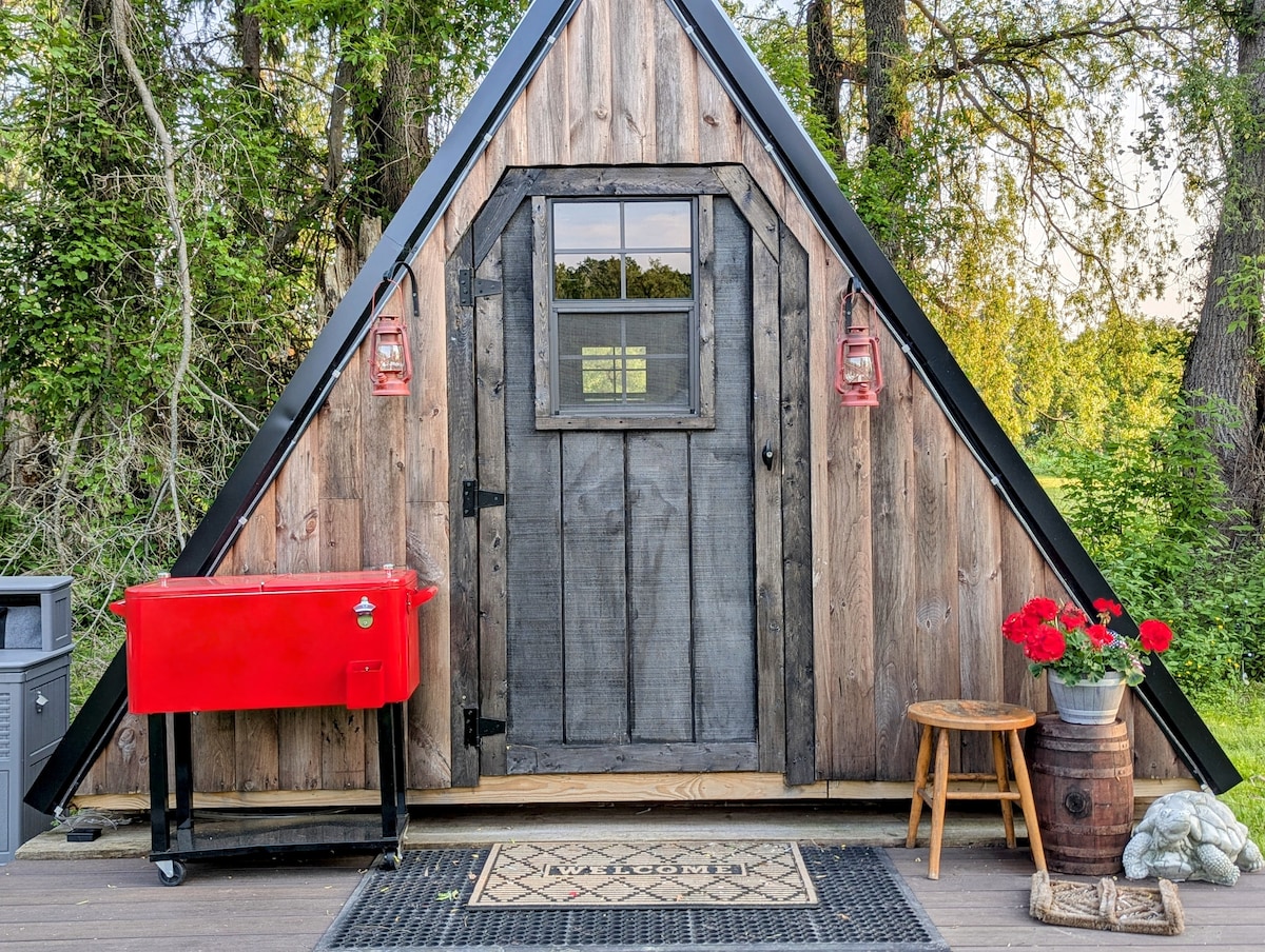 A quaint A-frame structure is presented with a rustic wooden exterior, complemented by a large front door and a window above. Red lanterns adorn either side of the entrance. Nearby, a red grill stands on a wooden deck, surrounded by vibrant flowers and a decorative welcome mat.