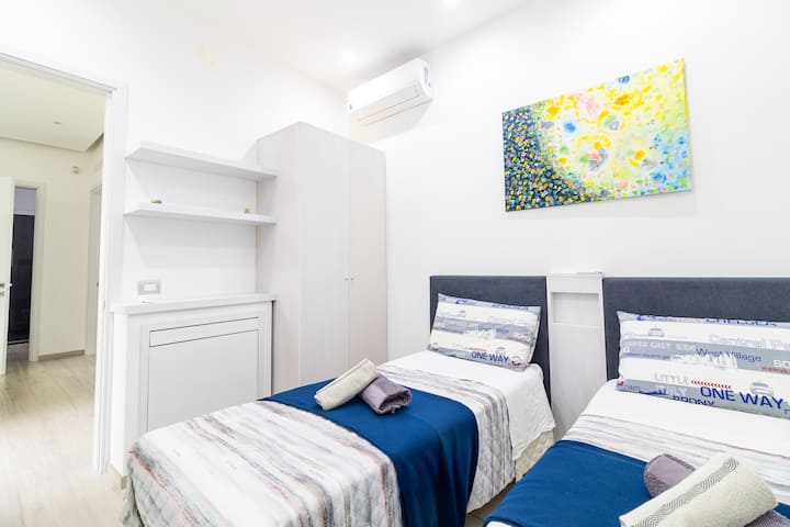 Dormitorio con dos camas individuales con cabecera gris, mantas azules y cubrecamas a rayas. Armario blanco grande, estantes, aire acondicionado.