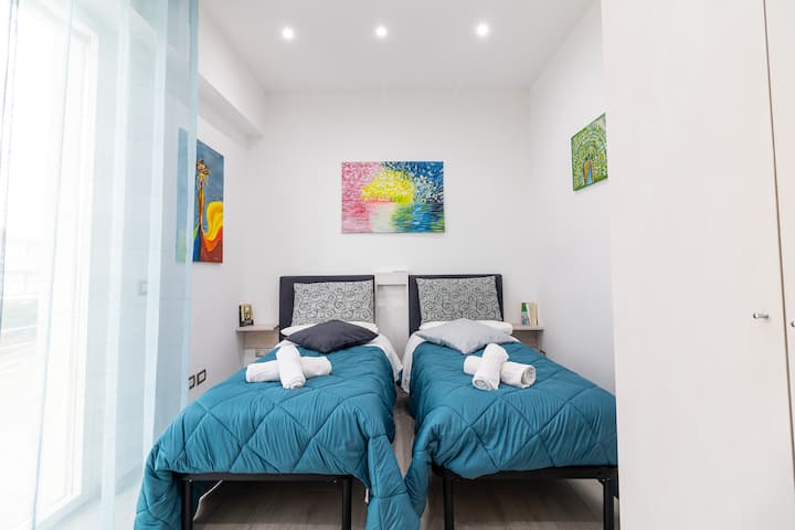 Habitación doble con ventana y cortinas transparentes de color azul claro. Armario blanco y dos camas individuales con mesitas de noche.