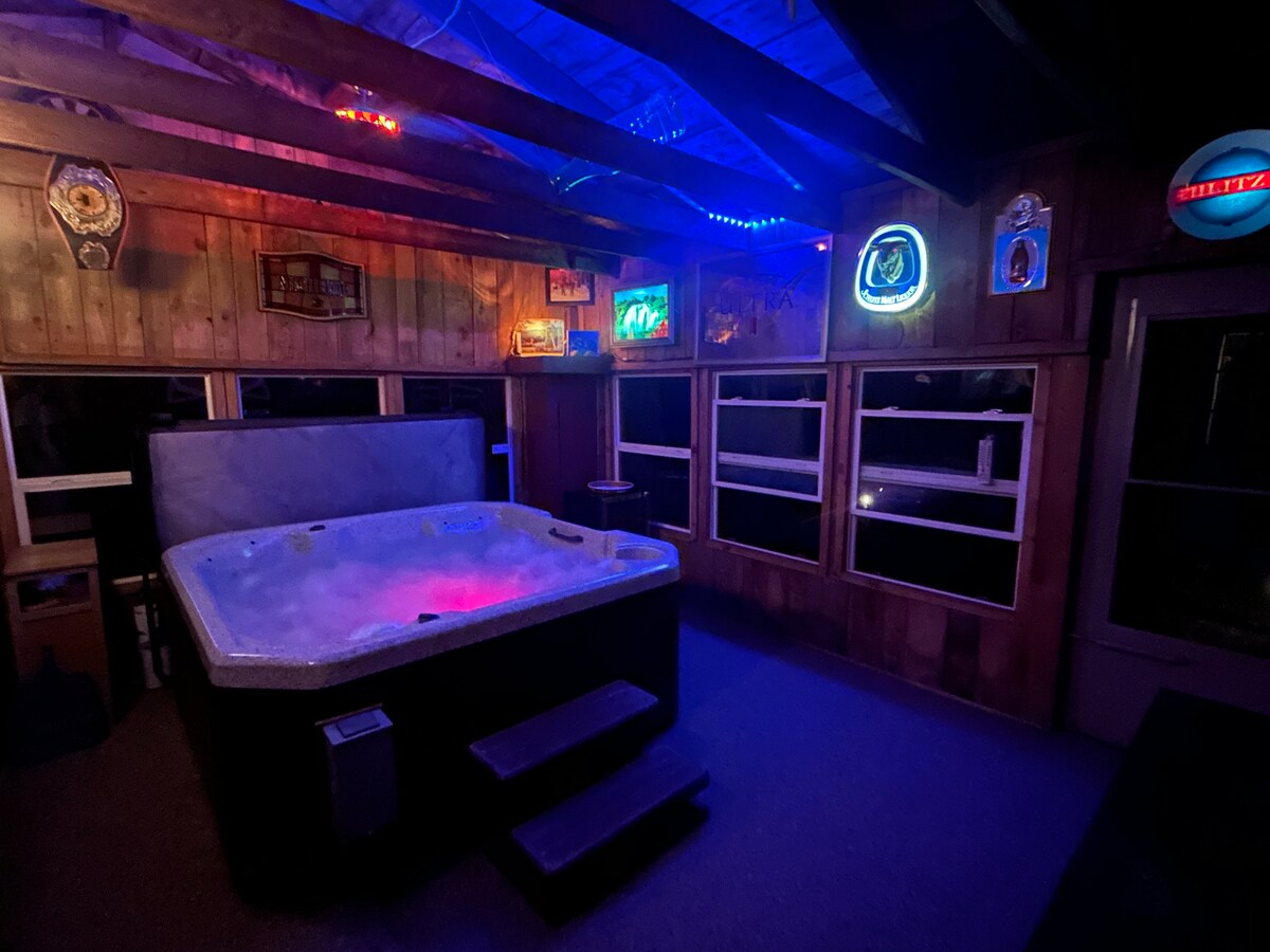 A hot tub is situated in a dimly lit room with wood paneling, featuring illuminated signs and soft lighting. Steps lead up to the tub, which is surrounded by large windows that provide glimpses of the outdoor scenery.