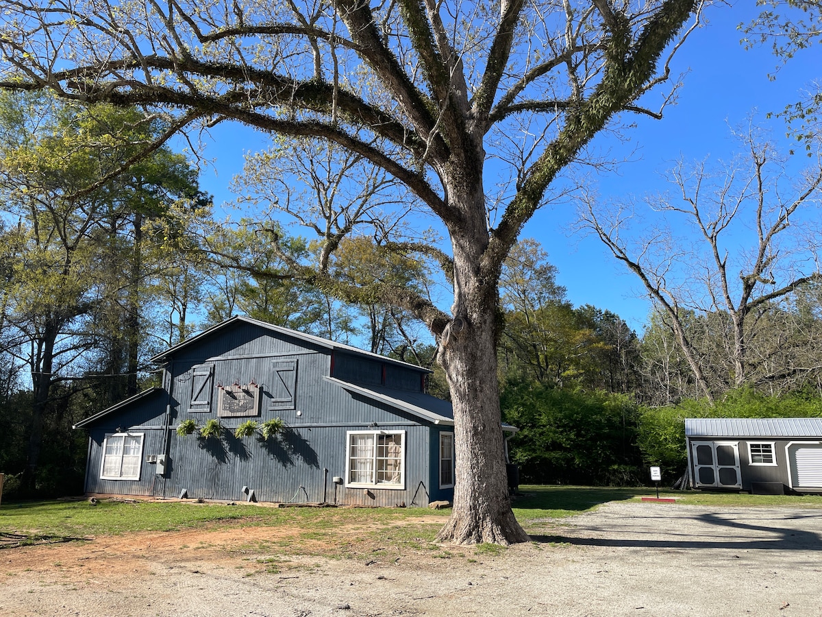 A rustic barn-style building is depicted in the image, surrounded by a well-maintained gravel area and large trees. The blue exterior contrasts with the green grass and clear blue sky, creating an inviting country setting.