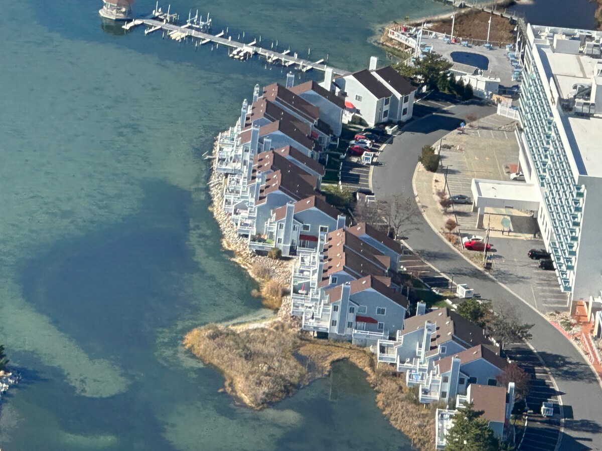 An aerial view captures a cluster of waterfront townhouses along the Assawoman Bay, characterized by light-colored exteriors and sloped roofs. A marina is visible nearby, with docks extending into the water. Surrounding landscaping includes patches of natural greenery and a paved parking area.