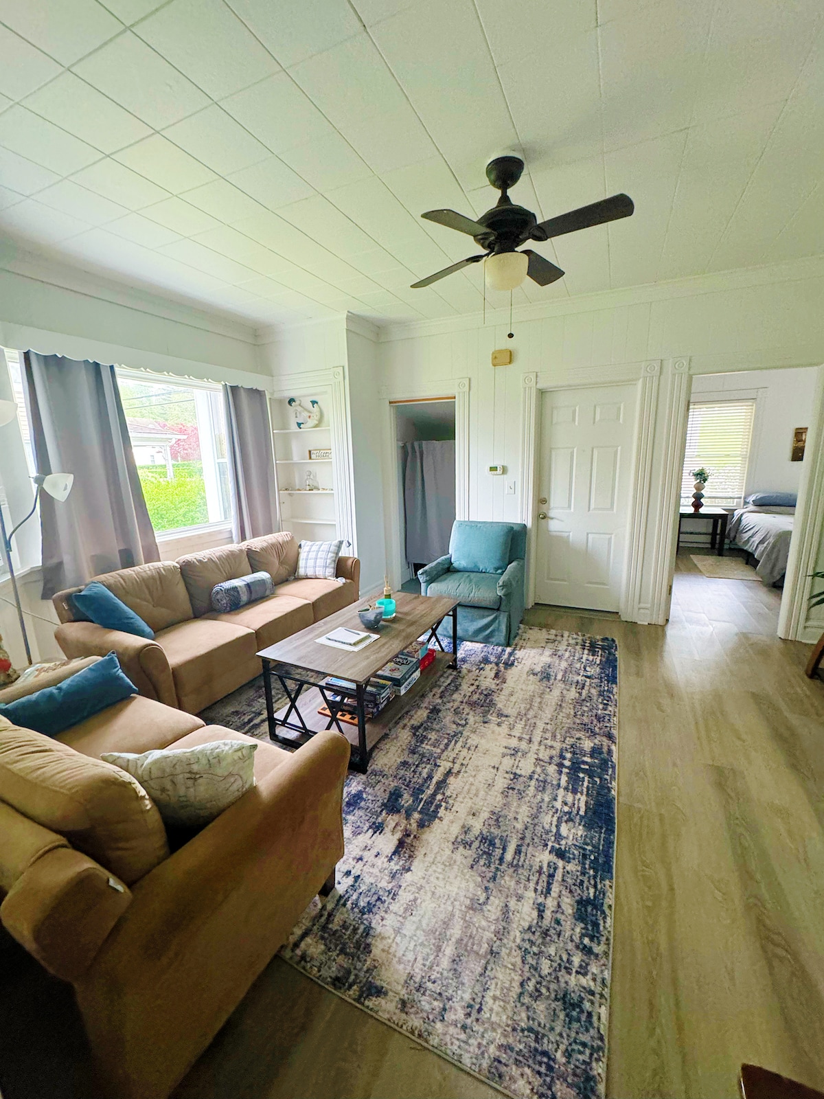 A comfortable living room area is presented, featuring two sofas and a chair arranged around a coffee table. Natural light enters through large windows framed by light curtains. A decorative rug covers part of the floor, directing attention towards an open doorway leading to a bedroom.