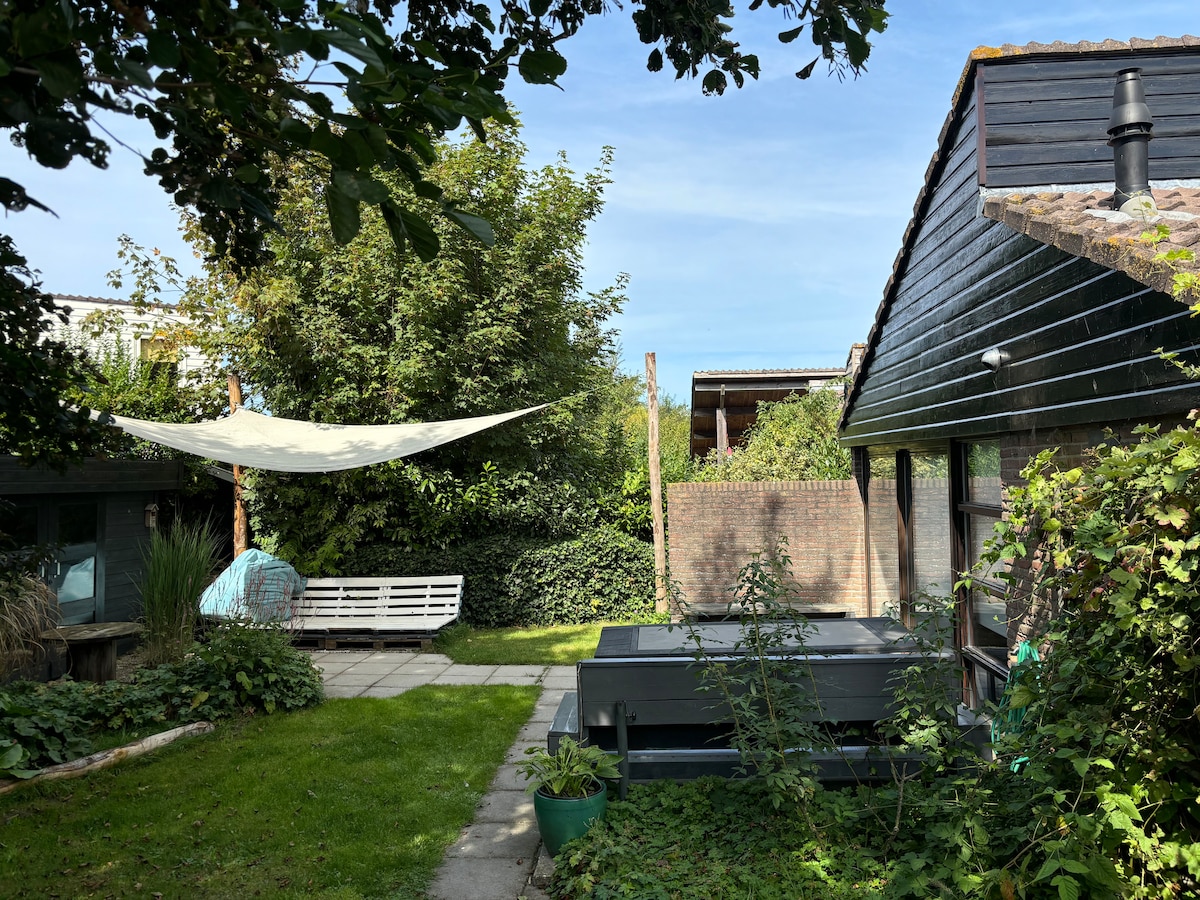 A serene outdoor space is surrounded by lush greenery, featuring a shaded area with a sail cloth installed overhead. A hot tub sits on stone paving adjacent to the house, while comfortable seating is placed under a tree, creating a relaxed atmosphere.