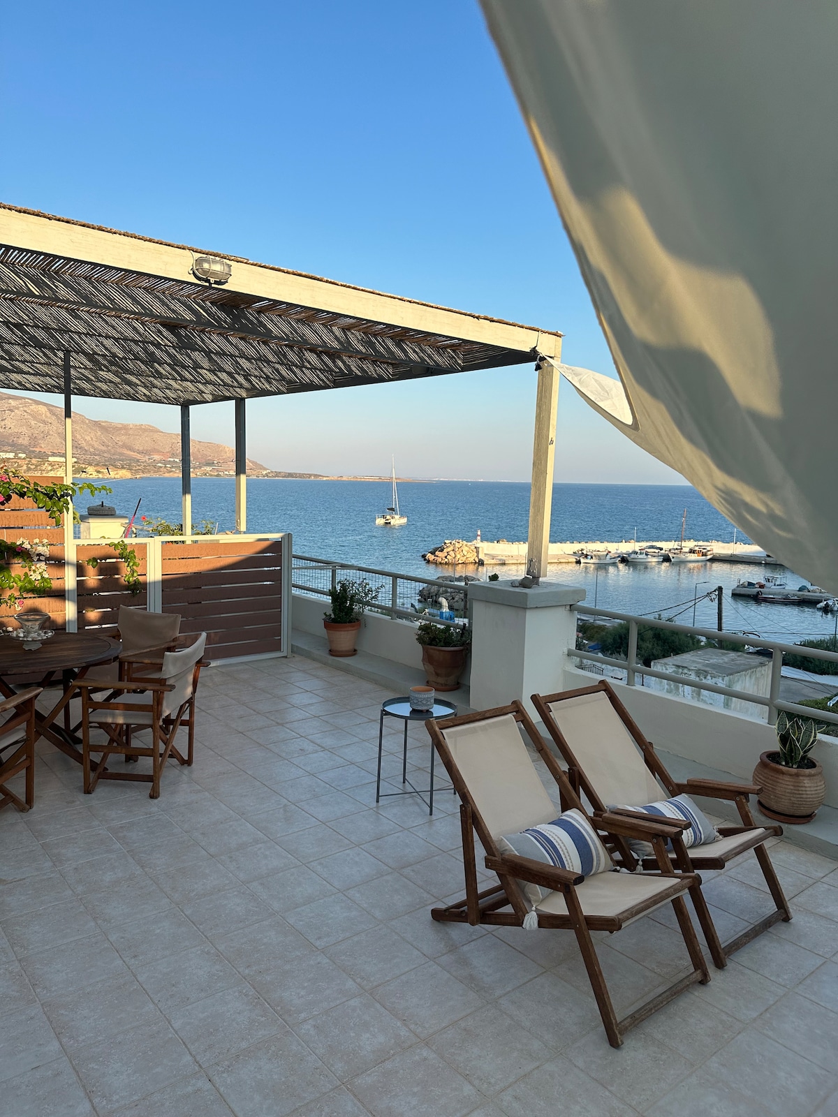 A sunlit terrace offers seating for relaxation, featuring two wooden chairs with striped cushions. Potted plants add greenery, while a stunning view of the sea and the marina is visible in the background. A shaded area provides respite from direct sunlight.