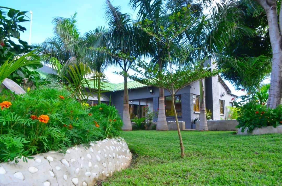 A spacious garden area is depicted, featuring well-maintained green grass and colorful flower beds. Lush palm trees are seen in the background, alongside a modern structure with large windows that invite natural light into the space.