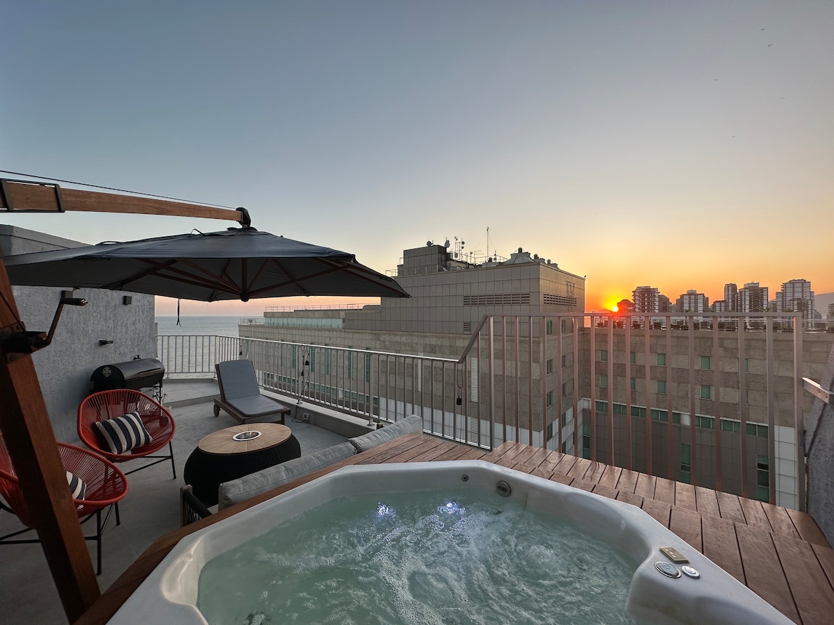 A rooftop area features a jacuzzi surrounded by wooden decking, with an umbrella providing shade. A comfortable seating arrangement includes bright red chairs and a small side table. The sunset casts warm hues across the sky, enhancing the view of the surrounding buildings and ocean.