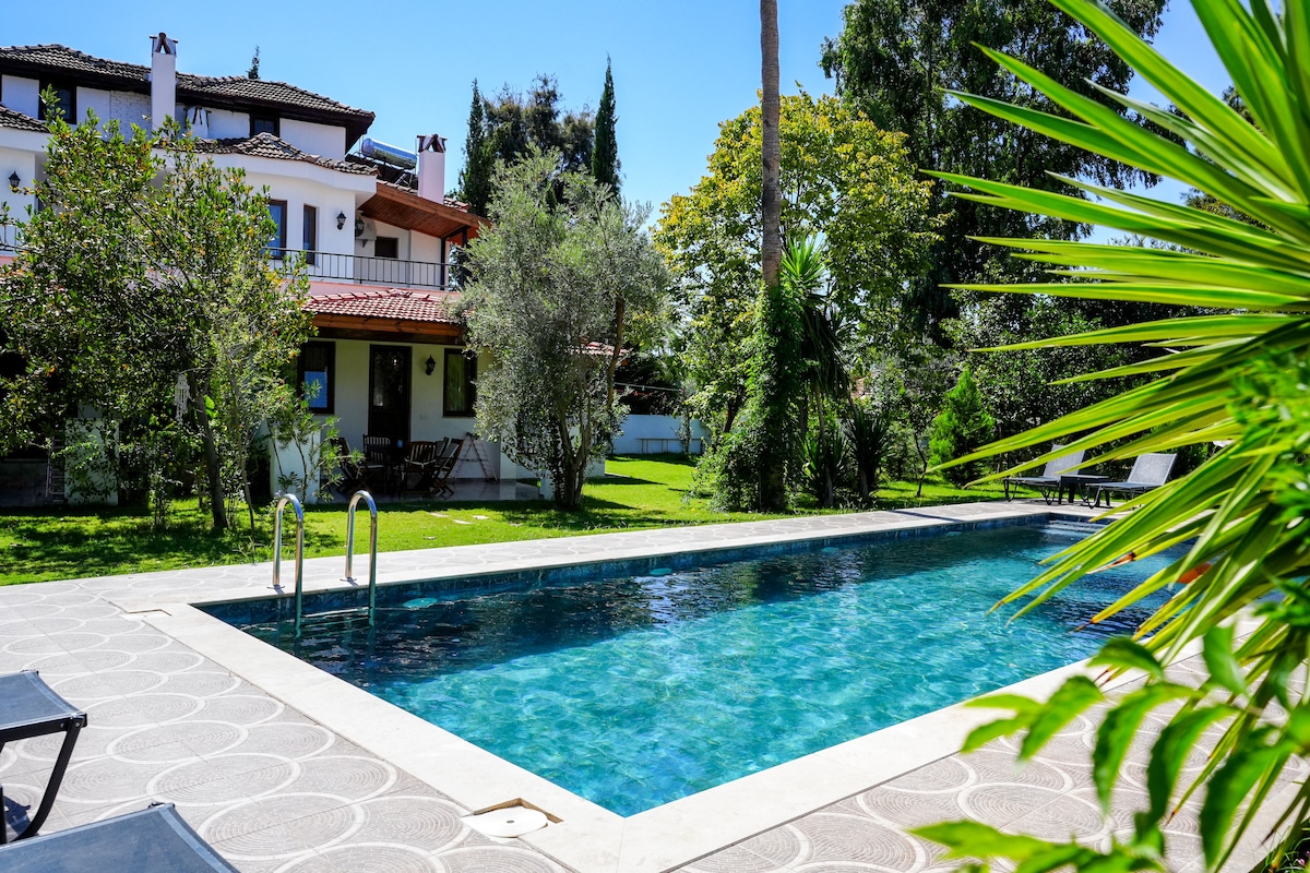 A large swimming pool is surrounded by a manicured garden featuring lush greenery and trees. Sun loungers are positioned beside the pool, offering spaces for relaxation. The two-story villa is visible in the background, highlighting its inviting outdoor areas.