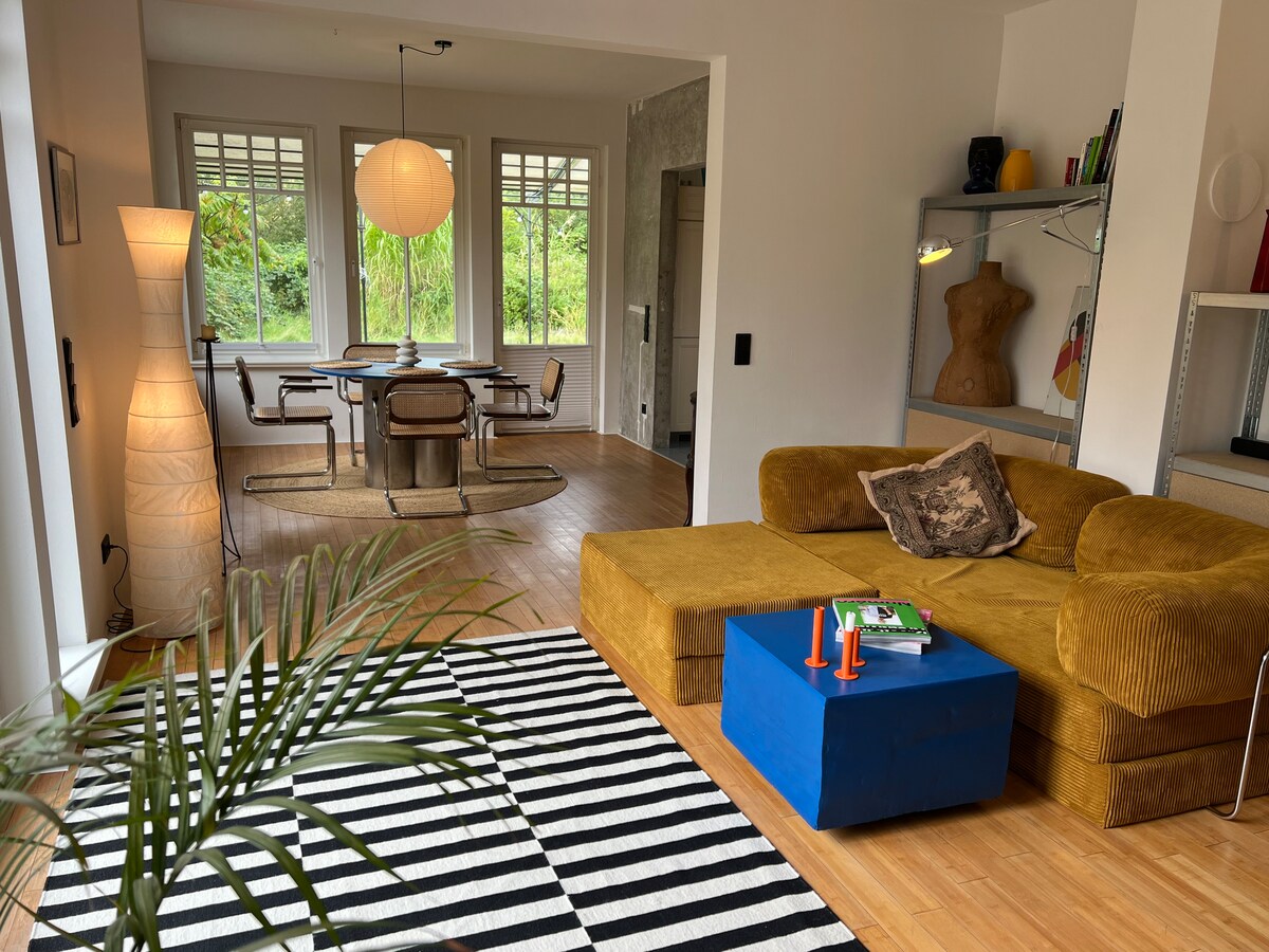 A stylish living area features a patterned rug with black and white stripes, complemented by a yellow sectional sofa. A blue coffee table is positioned centrally, while a round dining table with modern chairs is visible in the background, surrounded by large windows letting in natural light.