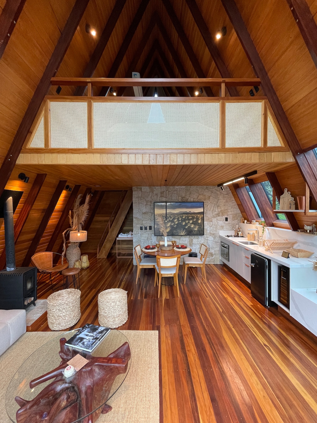 The interior features a spacious open-plan design with high A-frame ceilings. A cozy seating area is complemented by a modern kitchen and a round dining table for four. Natural wood finishes and stone accents create a warm atmosphere, with light streaming through large windows.