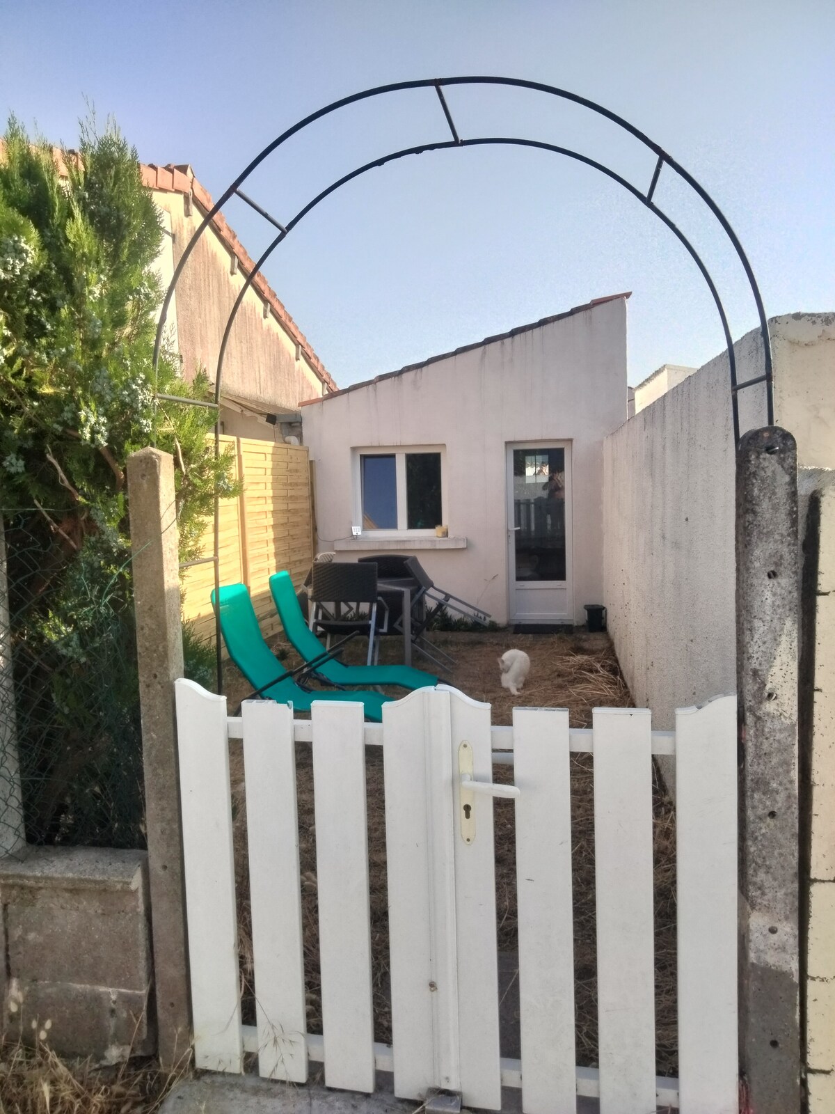 An entrance gate leads into a small garden area featuring two green lounge chairs positioned beside a table. A white building with a window is visible in the background, and an arched structure creates a focal point above the entryway.