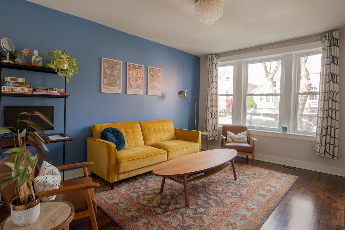 A cozy living area is highlighted by a bright yellow sofa facing a wooden coffee table atop a patterned rug. Three framed artworks adorn the blue wall, while natural light filters through two large windows with patterned curtains. A stylish plant adds a touch of greenery.