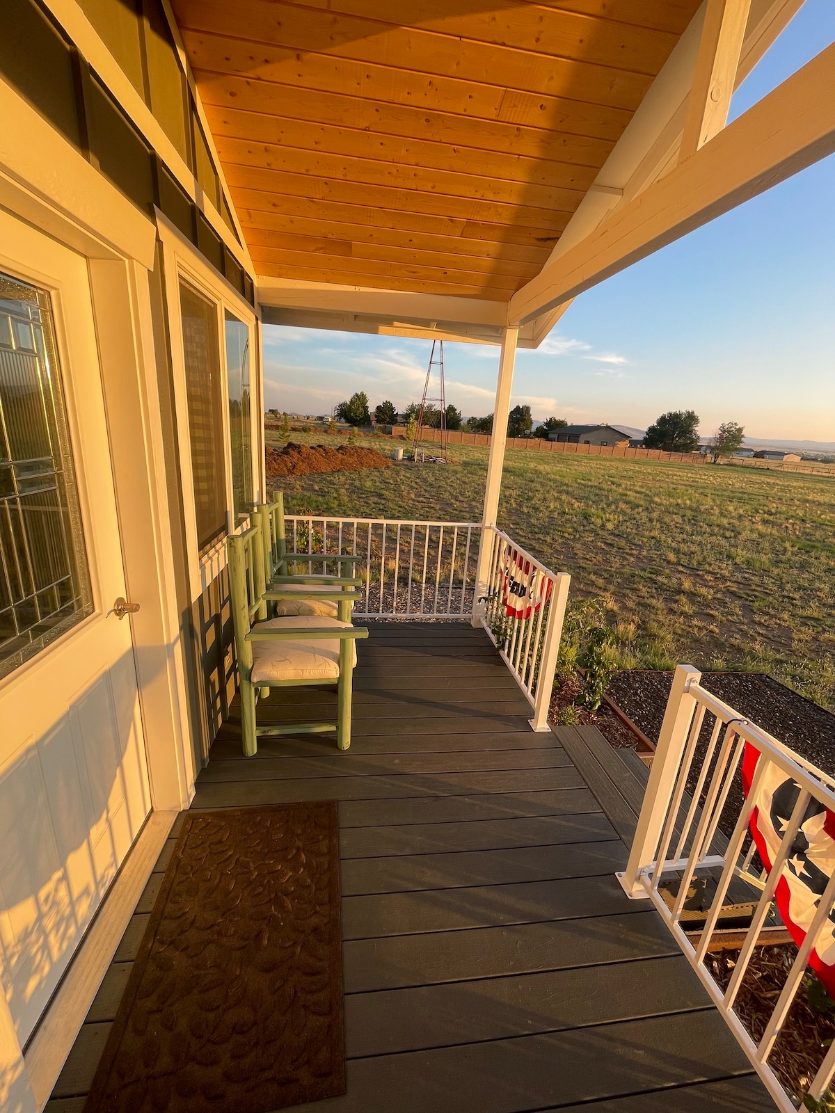A private porch is shown with a green chair positioned beside the entrance, providing an inviting spot to relax. The scene features open fields in the background, enhanced by a sunset sky, while the railing adds a touch of charm to the outdoor space.