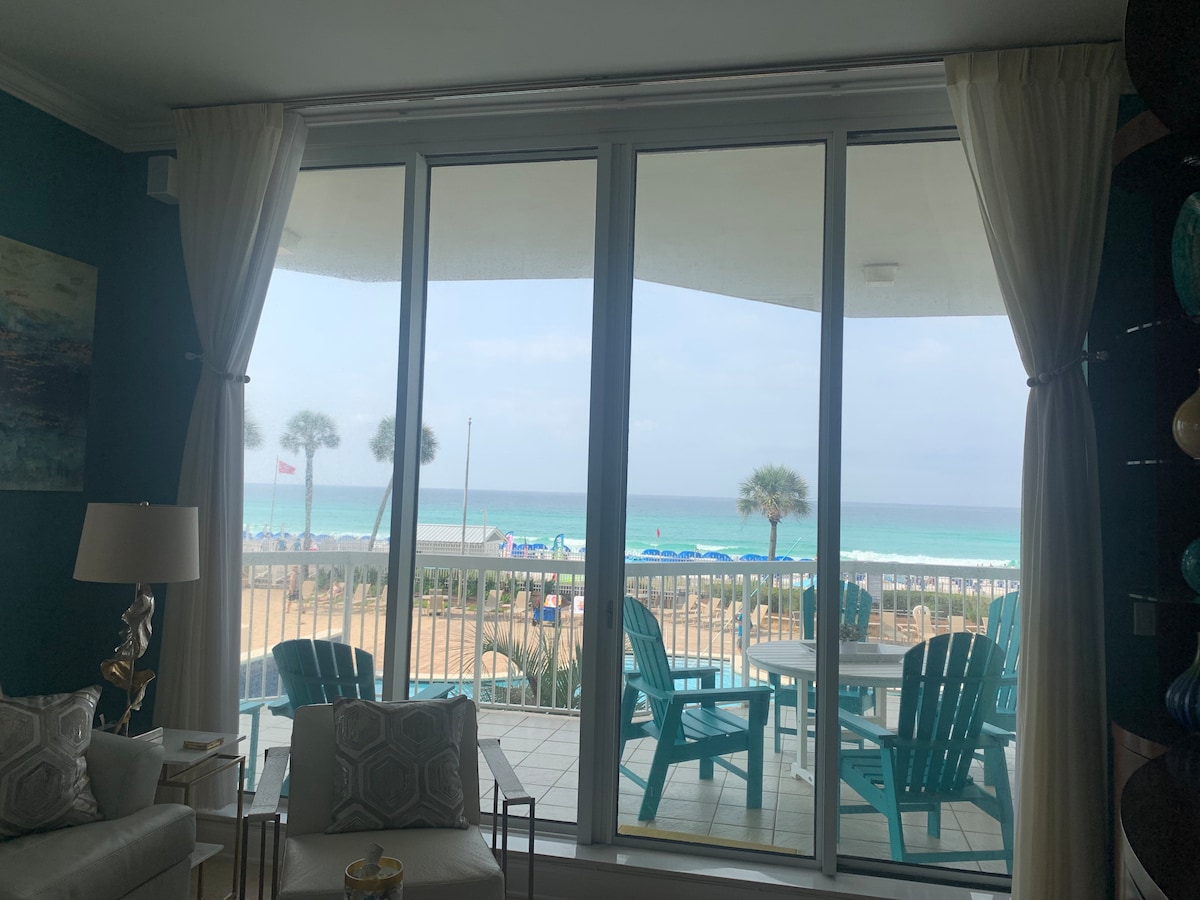 A large glass sliding door offers unobstructed views of the beach and ocean. Beyond the terrace, turquoise-blue outdoor chairs are positioned around a table, inviting relaxation. Palm trees frame the scene, while light coastal tones enhance the serene atmosphere.