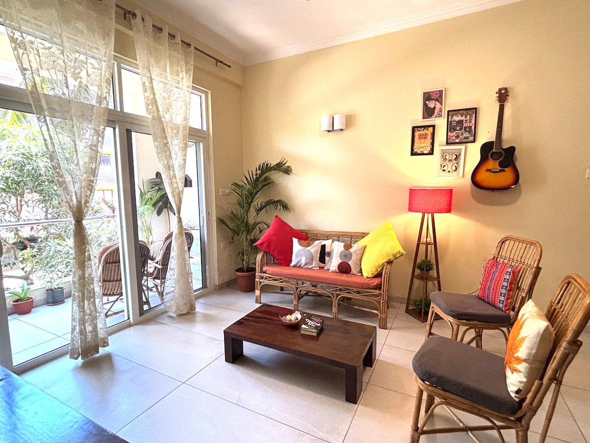 The living room features a combination of rattan furniture, including a sofa and three chairs, arranged around a wooden coffee table. Natural light enters through French sliding windows, illuminating the space. Decorative pillows of various colors and a guitar on the wall add visual interest, complemented by potted plants.